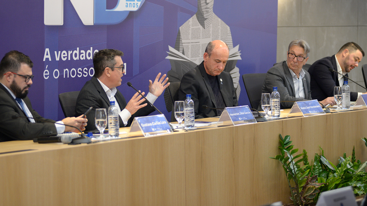 Foto horizontal de uma mesa de debates com cinco participantes sentados lado a lado. Entre eles, está o auditor fiscal de Controle Externo Hemerson, que aparece falando e gesticulando durante sua apresentação. Os debatedores estão posicionados atrás de uma bancada com microfones, placas de identificação e copos de água. Ao fundo, um painel institucional compõe o cenário do evento. 