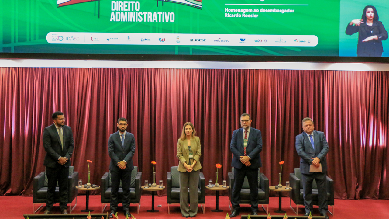Foto horizontal posada de cinco palestrantes, lado a lado, em pé, no palco do evento. Entre os presentes, Tatiana Hahn, Leonardo Cristóvam, Yuri Schneider, Fábio Santos e Marcelo Harger. O cenário é composto por tapete e cortinas bordôs, além de flores decorativas. No telão, na parte superior, o logo do congresso. 