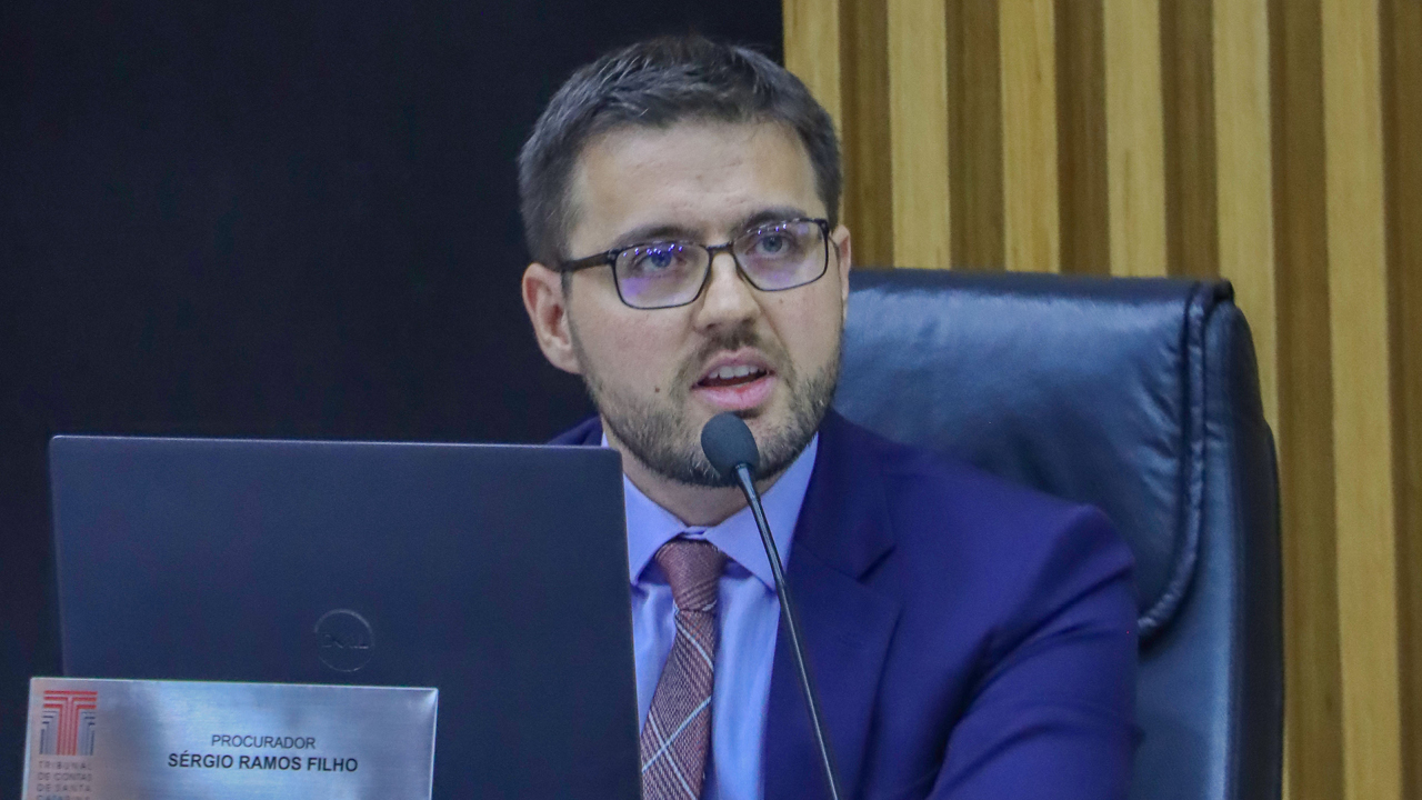 Foto horizontal do procurador-geral adjunto do MPTC/SC, Sérgio Ramos Filho, durante sessão. Ele é um homem branco, de cabelos e barba escuros. Veste terno e gravata e usa óculos de grau. Está sentado diante da bancada circular no Plenário. Tem expressão séria.