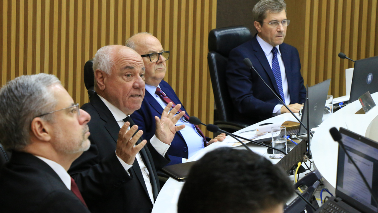 Foto do conselheiro Luiz Eduardo Cherem falando durante a sessão. É um homem branco, de cabelos grisalhos. Veste terno e gravata. Está com a expressão séria, sentado diante da bancada circular do Plenário. Ao seu lado, há outros conselheiros.