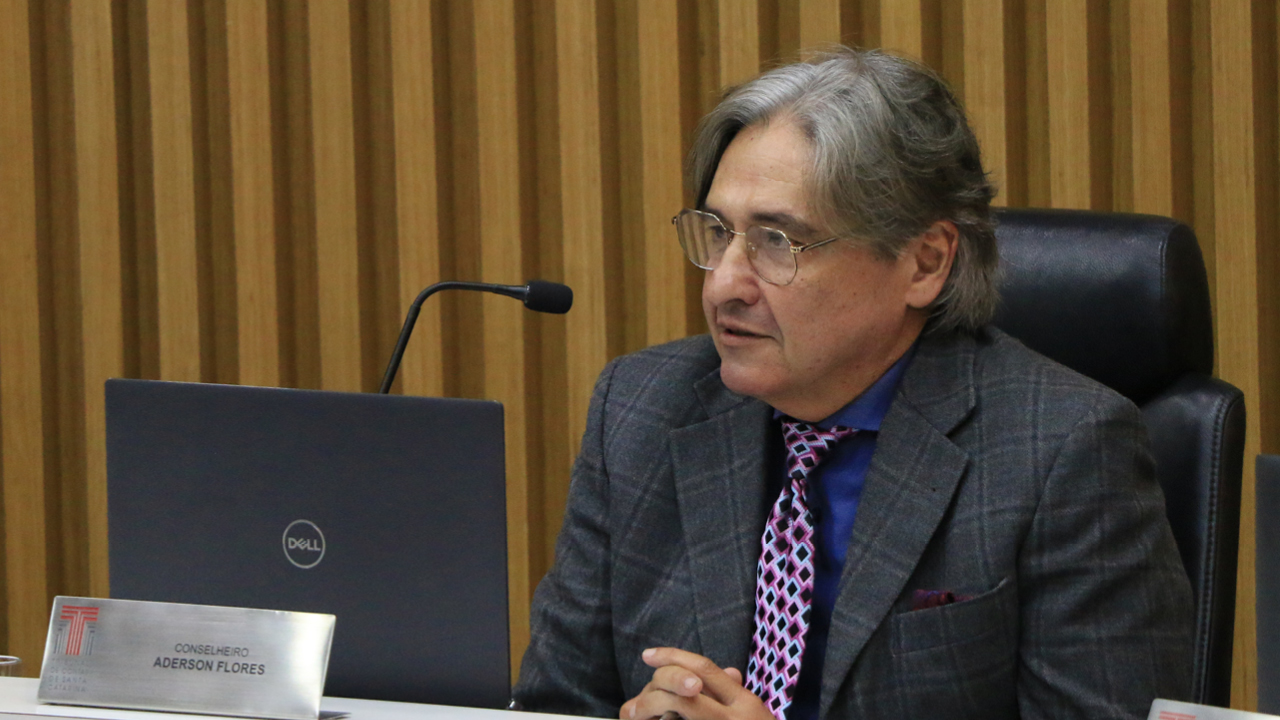 Foto do conselheiro Aderson Flores durante a sessão. É um homem branco, de cabelos grisalhos. Veste terno e gravata e usa óculos de grau. Está sentado diante da bancada circular no Plenário. Olha para a direita, com expressão séria.