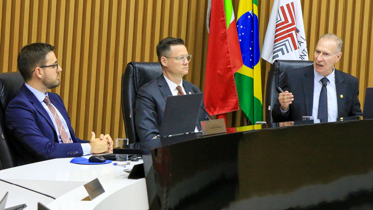 Foto horizontal do conselheiro Herneus De Nadal ao lado do secretário da Fazenda, Cleverson Siewert, e do procurador do MPTC/SC Sérgio Ramos Filho. Eles estão sentados diante da bancada circular do Plenário. O conselheiro Herneus fala ao microfone.