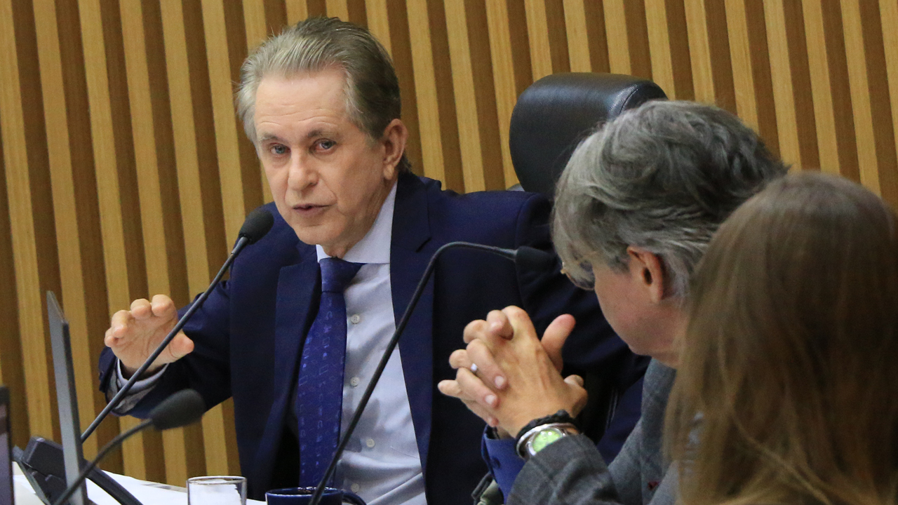 Foto horizontal do relator, conselheiro Luiz Roberto Herbst, falando ao microfone durante a sessão. Ele é um homem branco, de cabelos grisalhos. Veste terno e gravata e está sentado diante da bancada circular do Plenário.