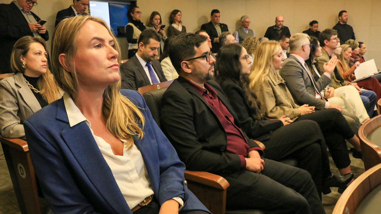 Foto da plateia acompanhando a sessão. Em primeiro plano, homens e mulheres sentados em poltronas observam atentamente a sessão com expressões concentradas. Destaca-se uma mulher de blazer azul e camisa branca na primeira fileira. Ao fundo, outras pessoas acompanham a sessão sentadas ou em pé junto à parede, algumas utilizando celulares.