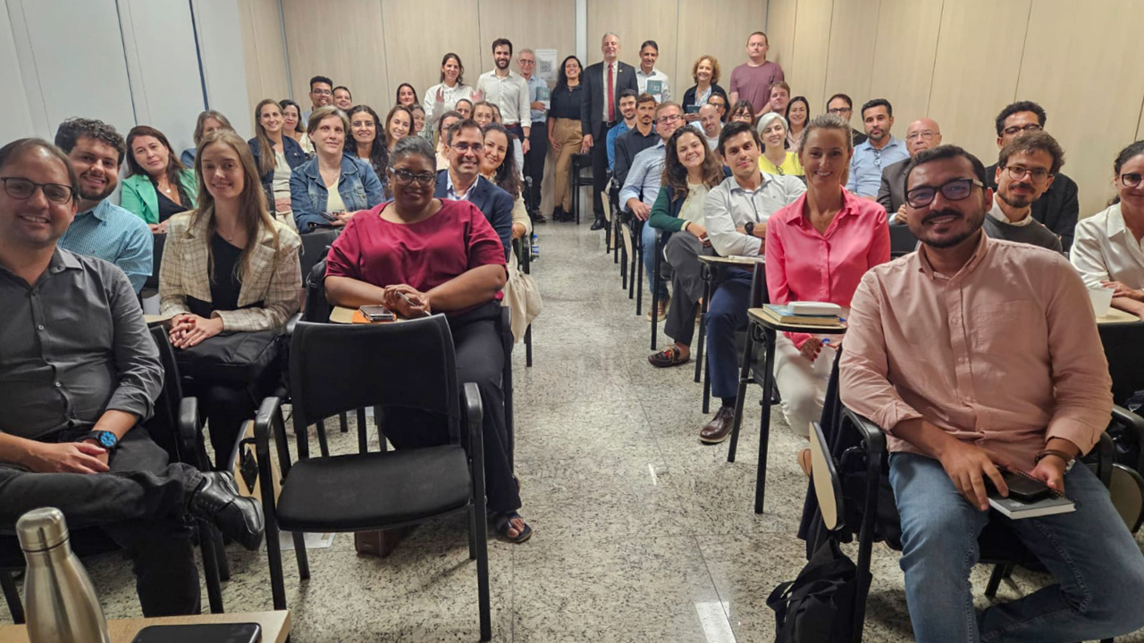 Imagem em ambiente de sala de capacitação, com os participantes do minicurso distribuídos em cadeiras e voltados para a câmera. Ao fundo da sala, aparecem os condutores da atividade e autoridades presentes no evento, reunidos em pé.  