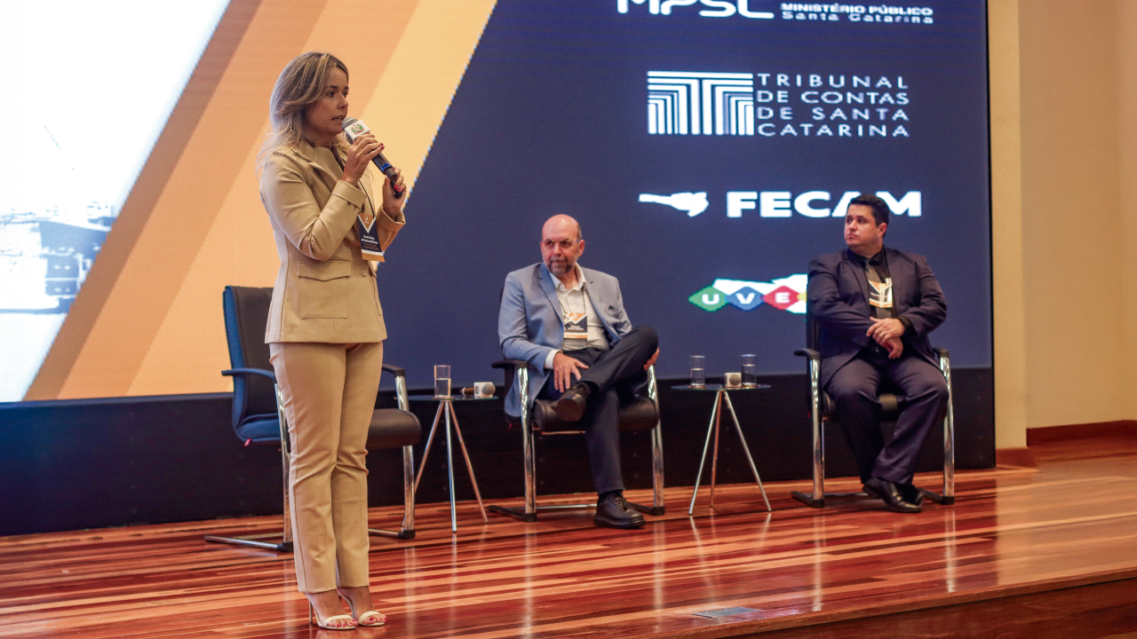 Foto da diretora da DGO, Gissele Nunes, no palco, palestrando. Ao fundo, sentados, o diretor do Icon, Moisés Hoegenn e o assessor da presidente Flávio Martins