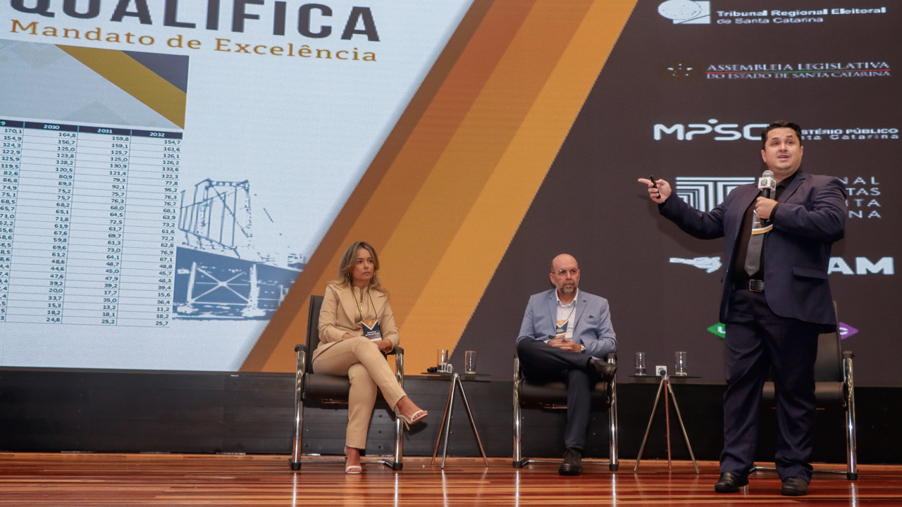 Foto do assessor da presidente, Flávio Martins, palestrando, no palco do auditório da Alesc. Ao fundo, a diretora da DGO, Gissele Nunes, e o diretor do Icon, Moisés Hoegenn
