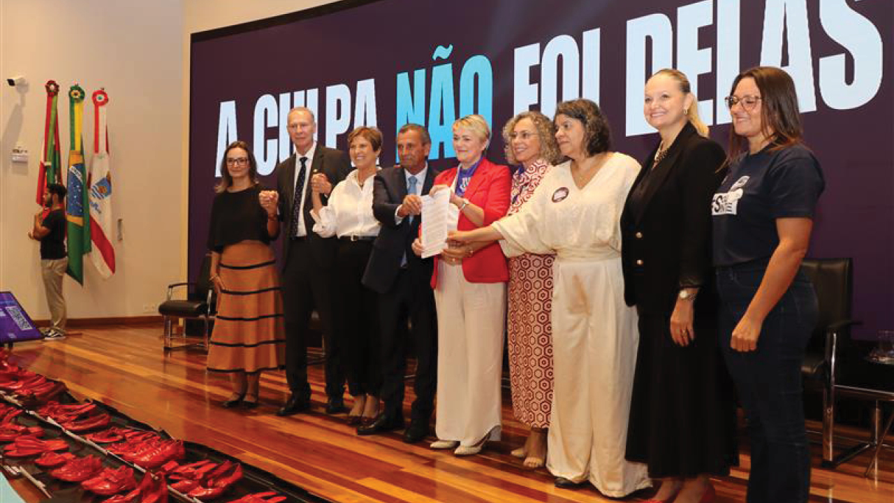 Foto horizontal das autoridades representantes das instituições participantes, alinhadas lado a lado para o registro oficial após a assinatura do acordo. Entre eles estão o presidente Herneus De Nadal, a deputada Luciane Carminatti, representantes do governo do estado e de outras entidades. Alguns seguram o documento assinado. Ao fundo, um grande painel exibe a frase “A culpa não foi delas”, reforçando a mensagem do evento.