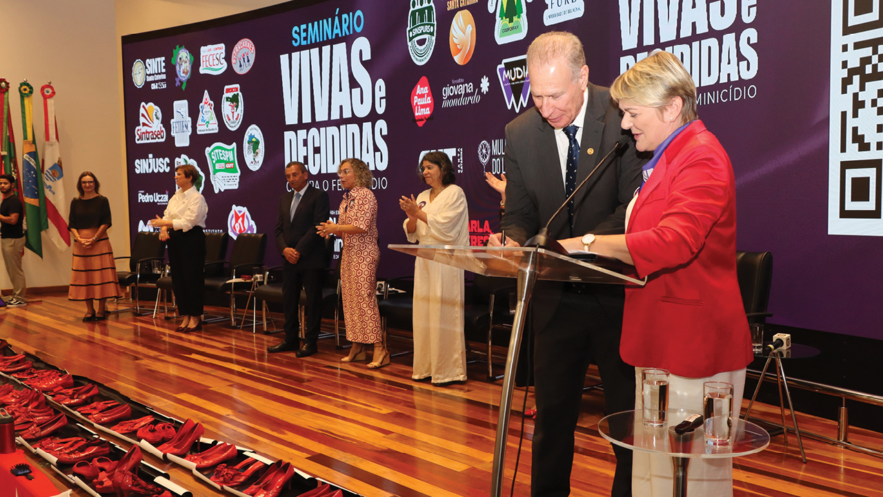 Banner horizontal com foto em auditório durante um evento institucional sobre enfrentamento à violência contra as mulheres. Em primeiro plano, o presidente Herneus De Nadal está ao lado da deputada Luciane Carminatti, ambos em pé diante de um púlpito transparente, assinando um documento. Ao fundo, outras autoridades estão alinhadas no palco, algumas aplaudindo o momento. A parede atrás do palco exibe um grande painel com o título “Seminário Vivas e Decididas” e diversos logotipos de instituições apoiadoras. 