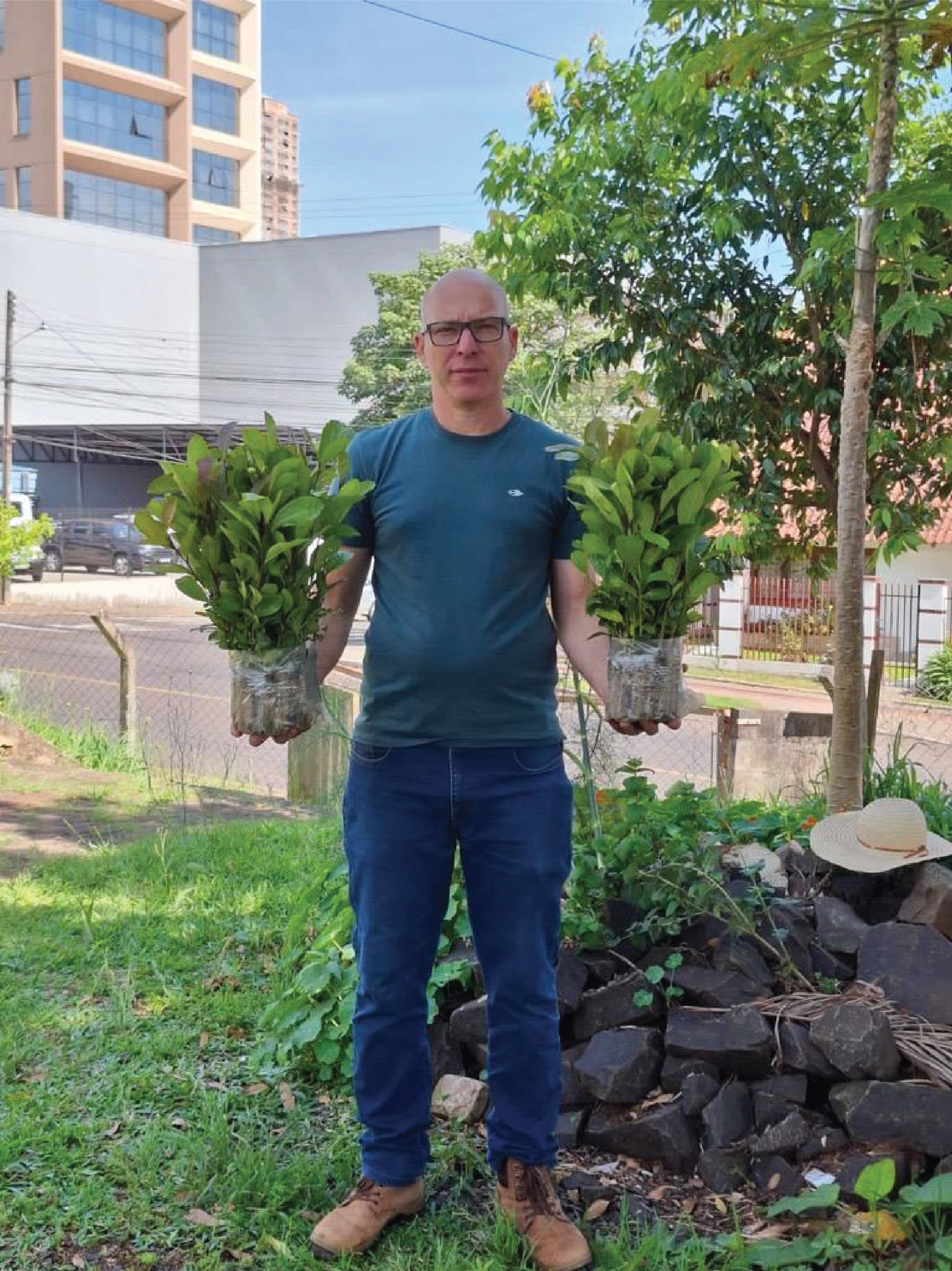 Foto da entrega realizada em Chapecó. Há um homem segurando duas mudas recebidas. Ele está em um espaço aberto, com vegetação e prédios atrás. 