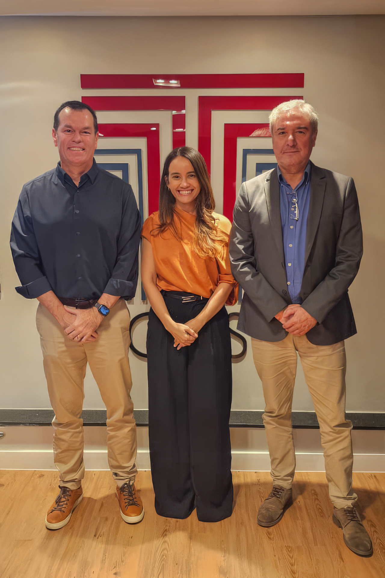 Foto vertical de Julia Maria Leal dos Santos ao centro, sorrindo, ao lado de seus orientadores: o conselheiro Adircélio de Moraes e o professor Luiz Henrique Cademartori. Os três estão em pé, em ambiente interno, com vestimentas sociais e fundo com plotagem do logo do TCE/SC na parede.