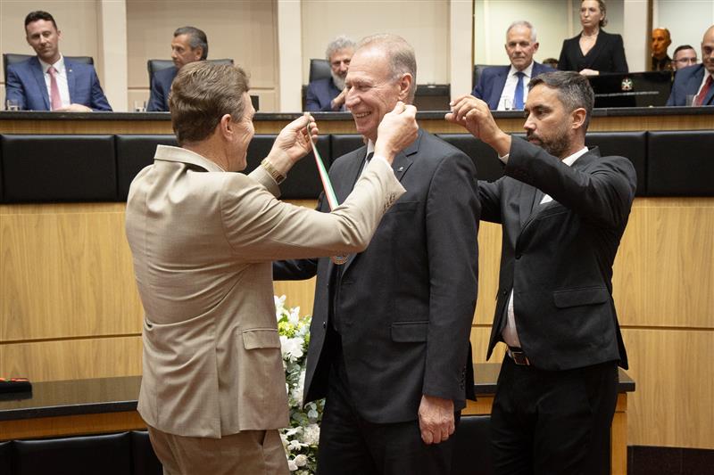 Foto do deputado Volnei Weber colocando a comenda no pescoço do conselheiro Herneus De Nadal, presidente do TCE/SC. Ao fundo, demais autoridades que compõem a bancada na Alesc. Herneus sorri.