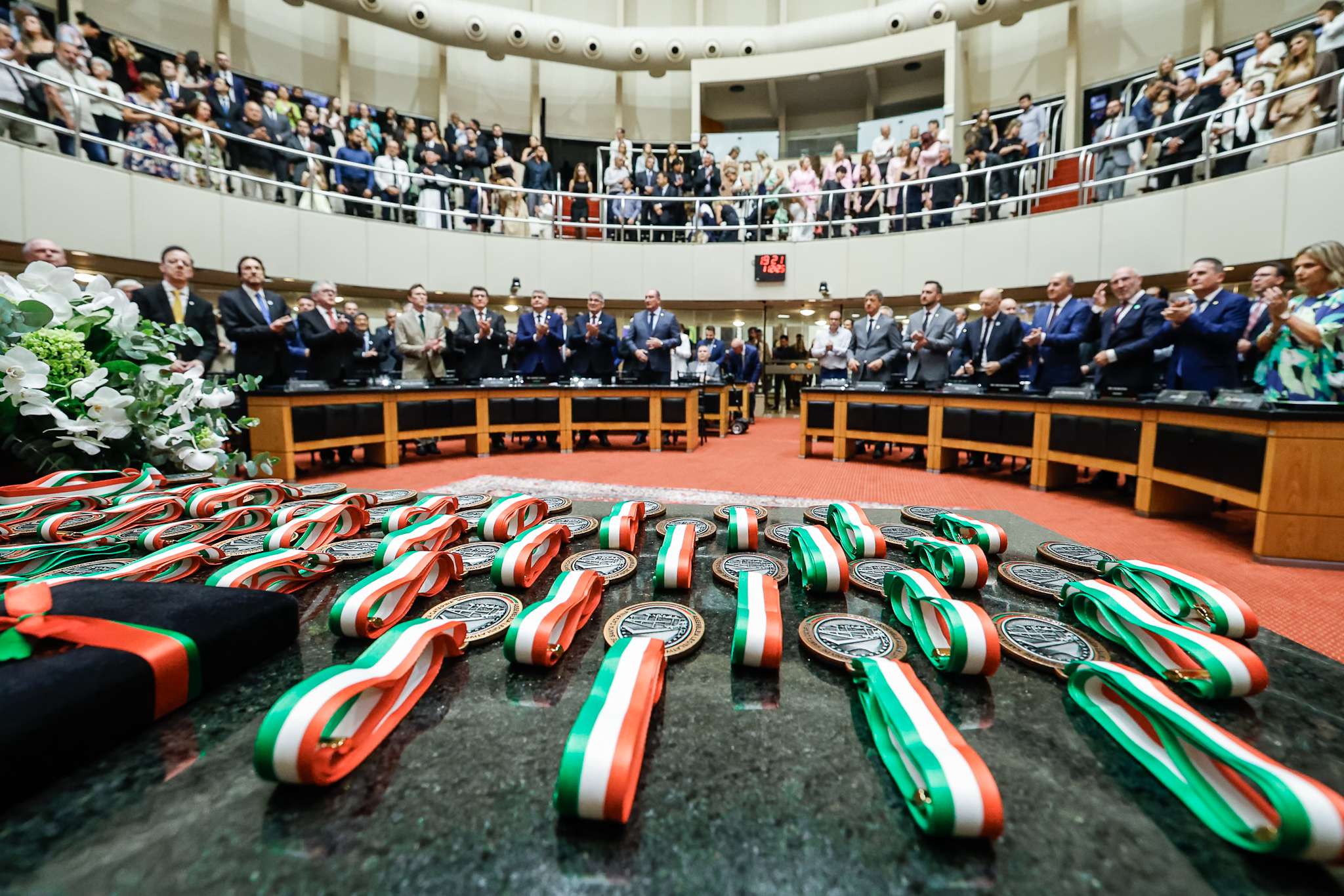 Foto horizontal de medalhas dispostas lado a lado sobre a bancada na Alesc. Ao fundo, aparecem várias pessoas que participam da sessão solene.