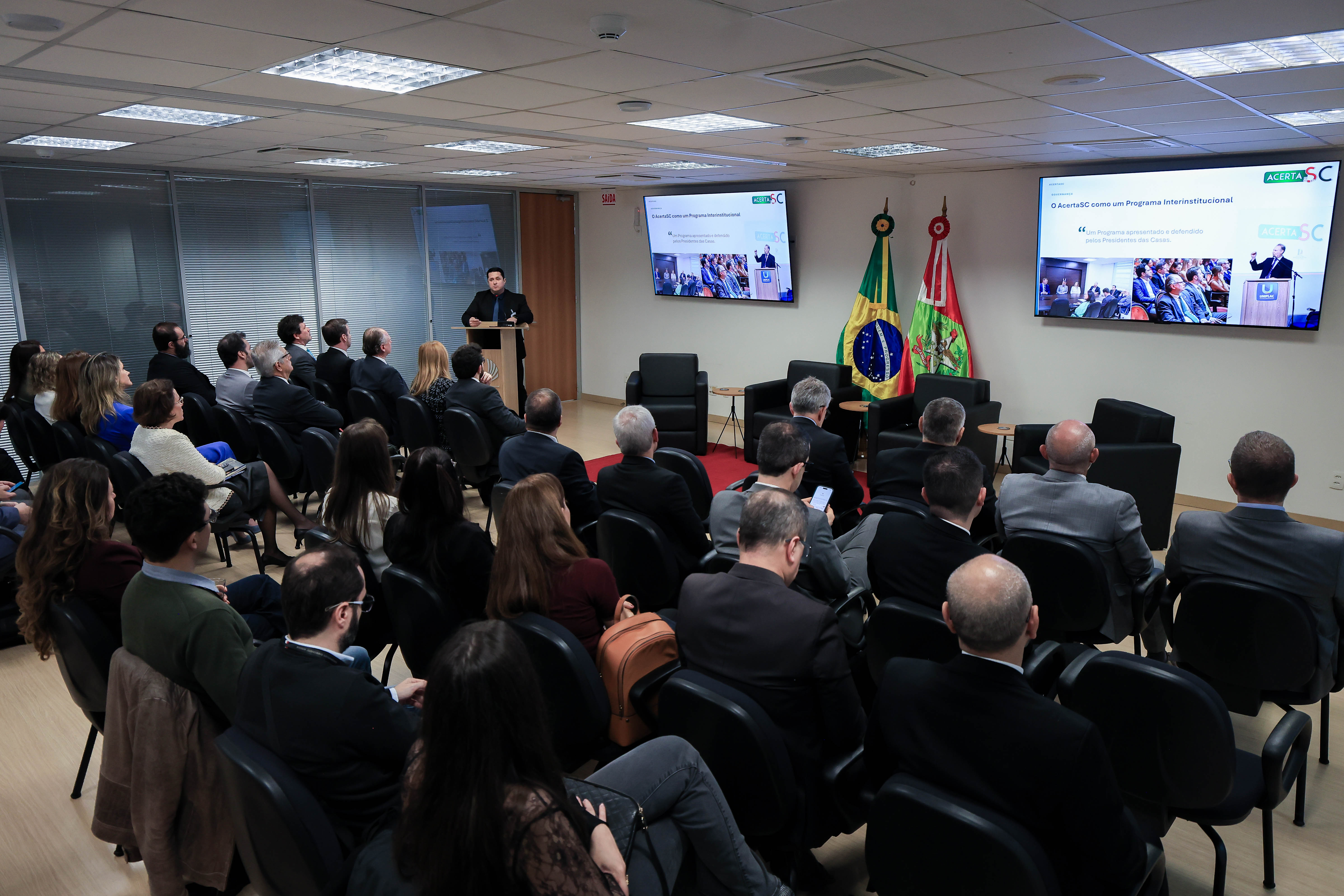 Foto ampla que mostra auditório cheio durante apresentação de Flávio Alves no TJSC. Há várias pessoas sentadas lado a lado, de costas. Ao fundo, no púlpito, Flávio fala ao microfone. Na parede, há dois televisores que exibem a apresentação.