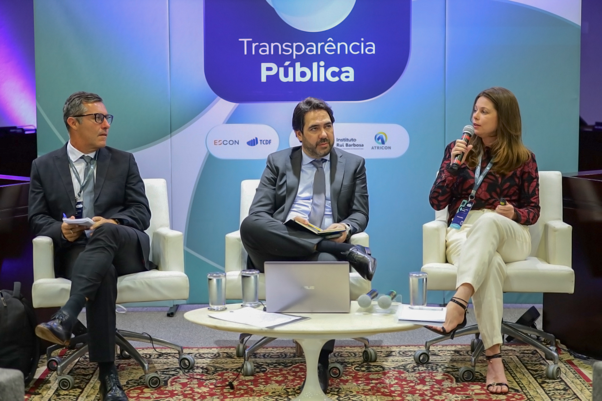 Foto horizontal que mostra a conselheira substituta Sabrina Nunes Iocken durante participação em painel, utilizando microfone enquanto se dirige ao público, ao lado de outros participantes sentados em poltronas, em ambiente institucional com painel ao fundo com o tema “Transparência Pública” e mesa de apoio com equipamentos e materiais.   