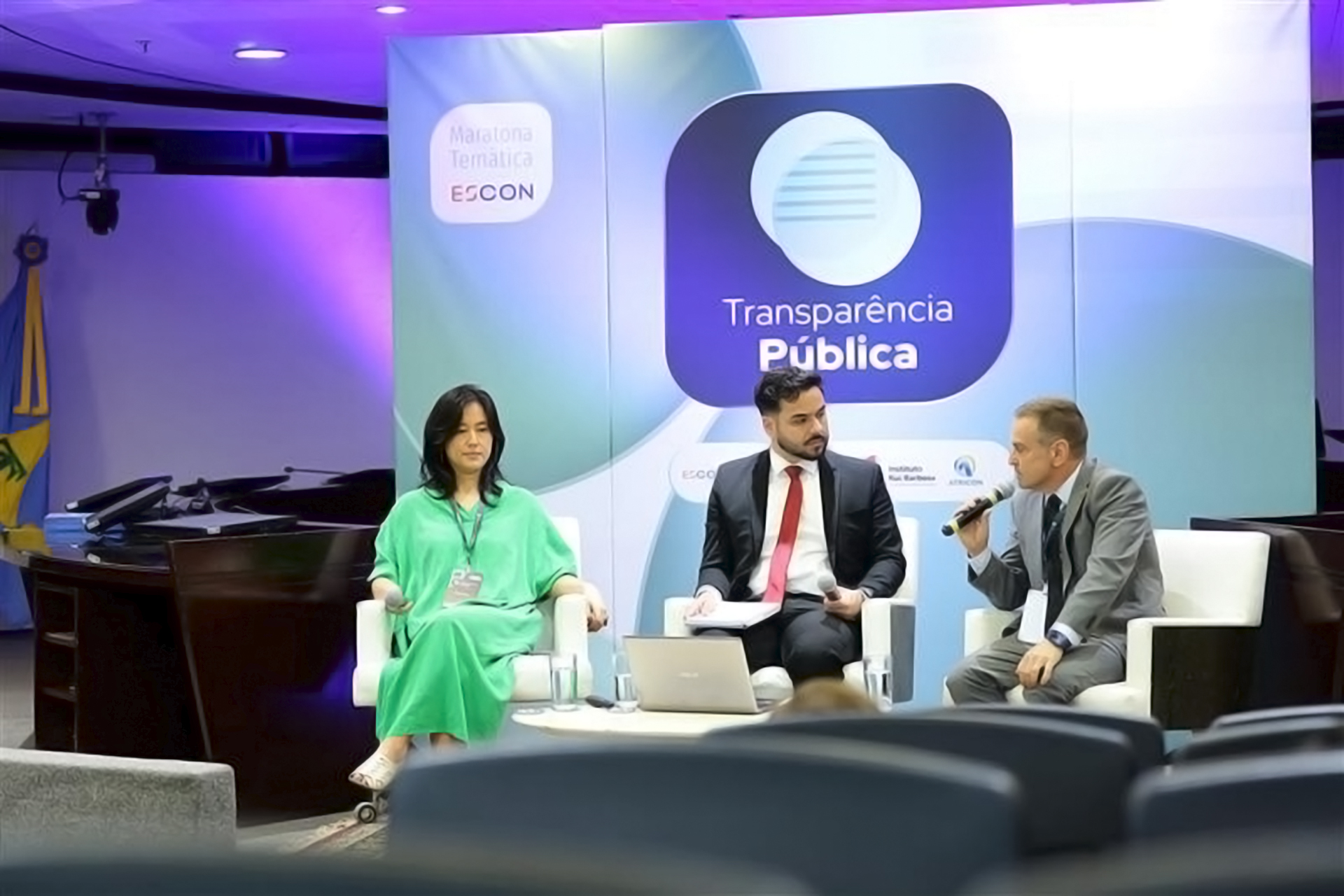 Foto horizontal do auditor fiscal de Controle Externo Renato Costa durante participação em painel, utilizando microfone enquanto se dirige ao público, ao lado de outros participantes sentados em poltronas, em ambiente institucional com painel ao fundo com o tema “Transparência Pública”. 