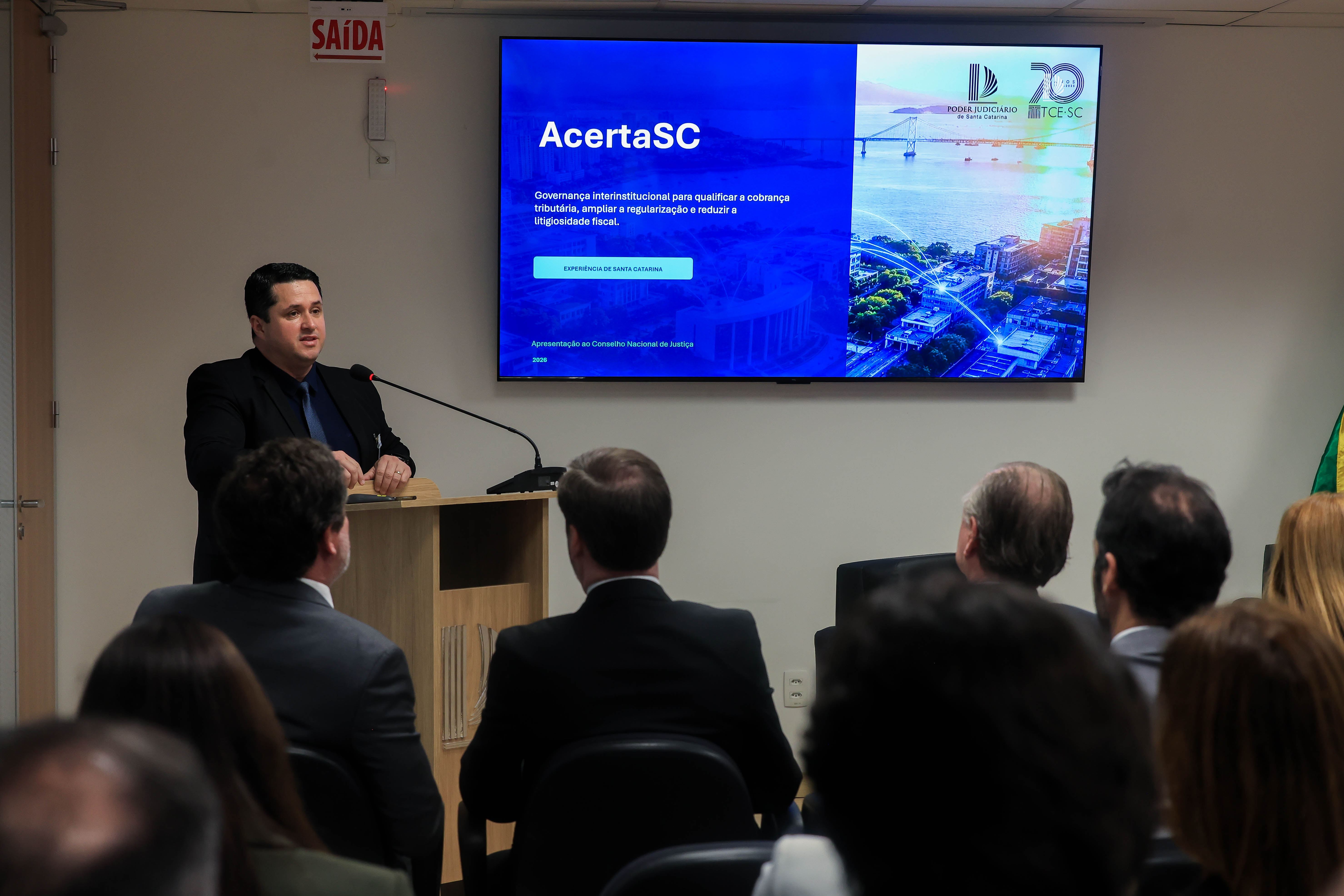 Foto horizontal do assessor Flávio Alves falando ao microfone no púlpito. Ao fundo, televisor exibe a apresentação sobre o Acerta SC.