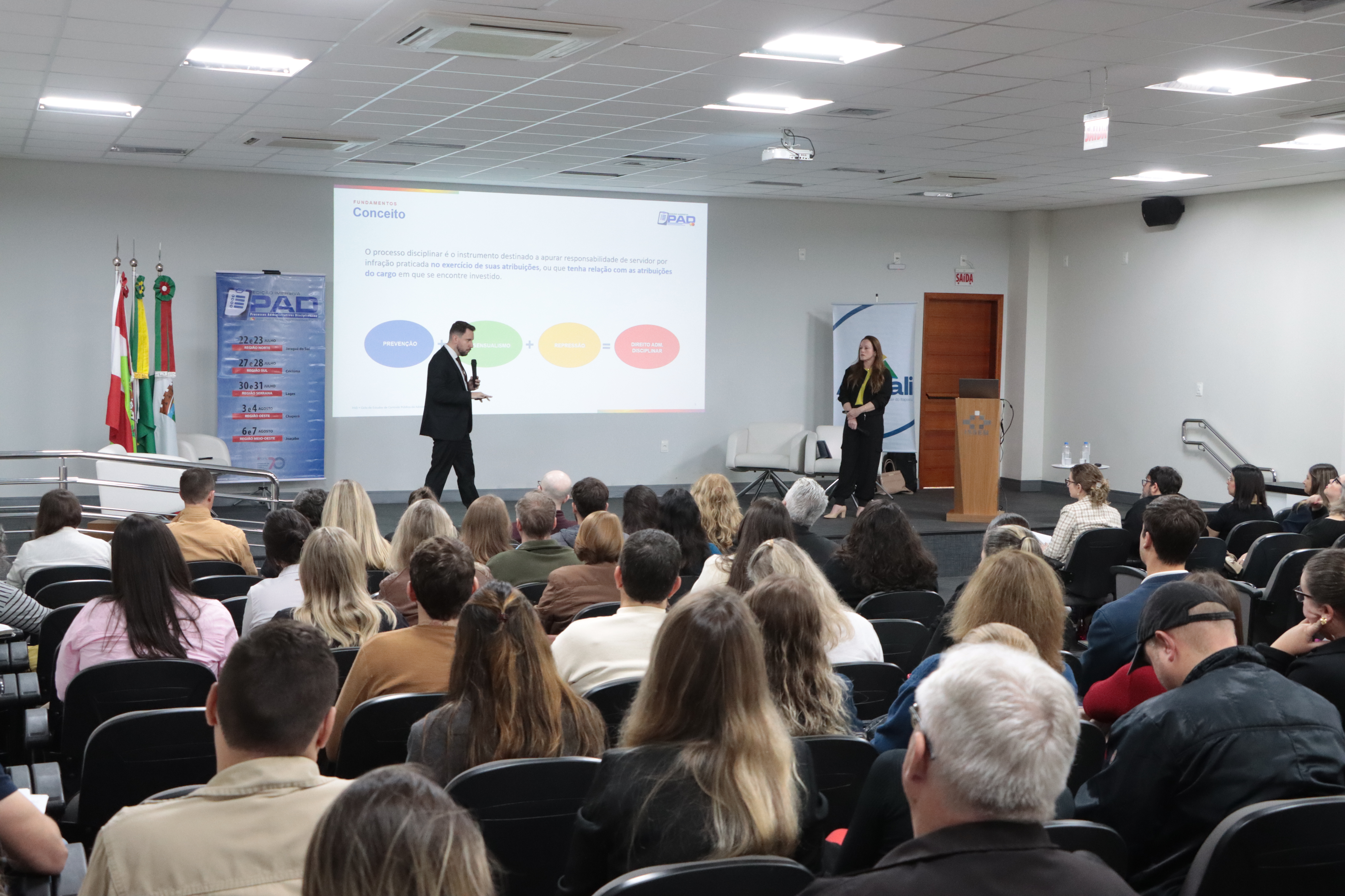 Foto horizontal que mostra um auditório lotado. Há várias pessoas sentadas em poltronas, lado a lado, na plateia, de costas. Ao fundo, no palco, os auditores Geovane Eziel e Vivian Savedra, palestrando. Eles estão em pé. Ao fundo, no telão, projeção de uma apresentação.