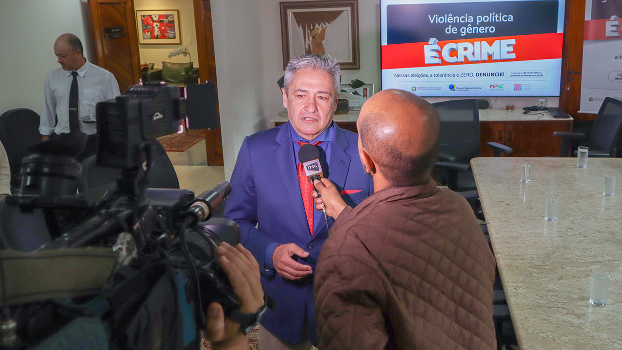 Foto do conselheiro Aderson Flores dando entrevista durante o lançamento da campanha. Ele está em pé, falando ao microfone, em frente às câmeras, com um repórter que o acompanha. Atrás deles, há um banner com a identidade visual da campanha. 