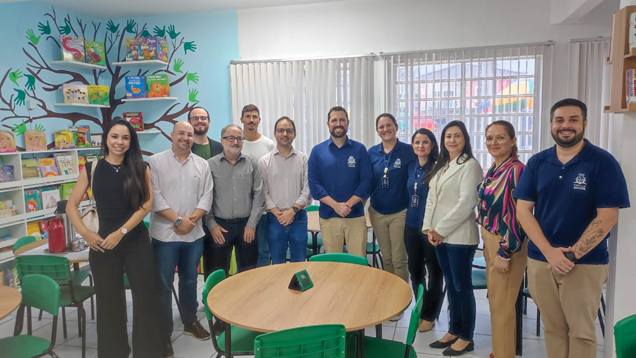 Foto posada do grupo em uma sala com várias estantes de livros ao fundo. Há doze pessoas em pé, uma ao lado da outra. Entre os presentes, os servidores do TCE/SC e membros da educação de Joinville. 