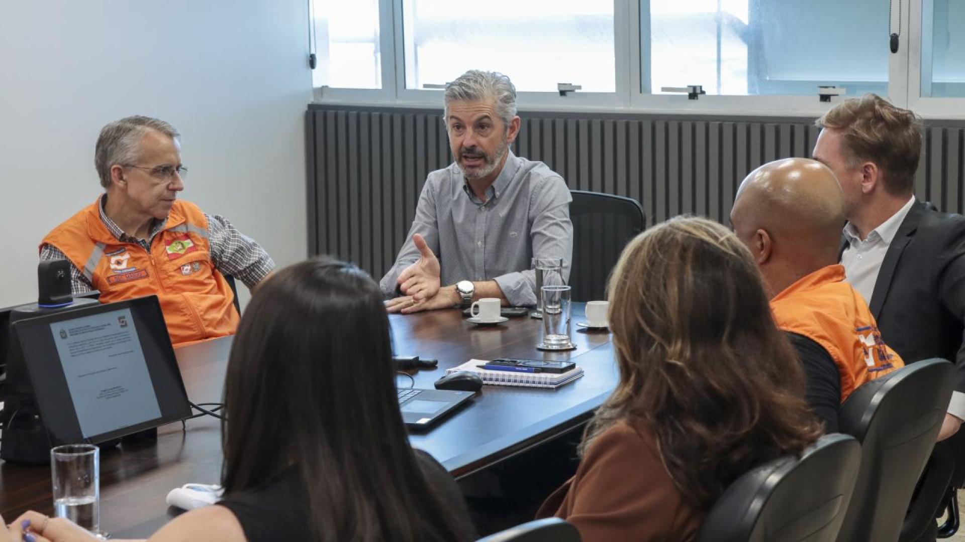 Imagem mostra seis pessoas sentadas ao redor de uma mesa. Quatro delas estão de costas. Aparecem de frente dois homens. À esquerda, um homem com cabelos grisalhos vestindo um colete alaranjado. Mais ao centro, um homem de cabelos grisalhos e barba vestindo uma camisa cinza