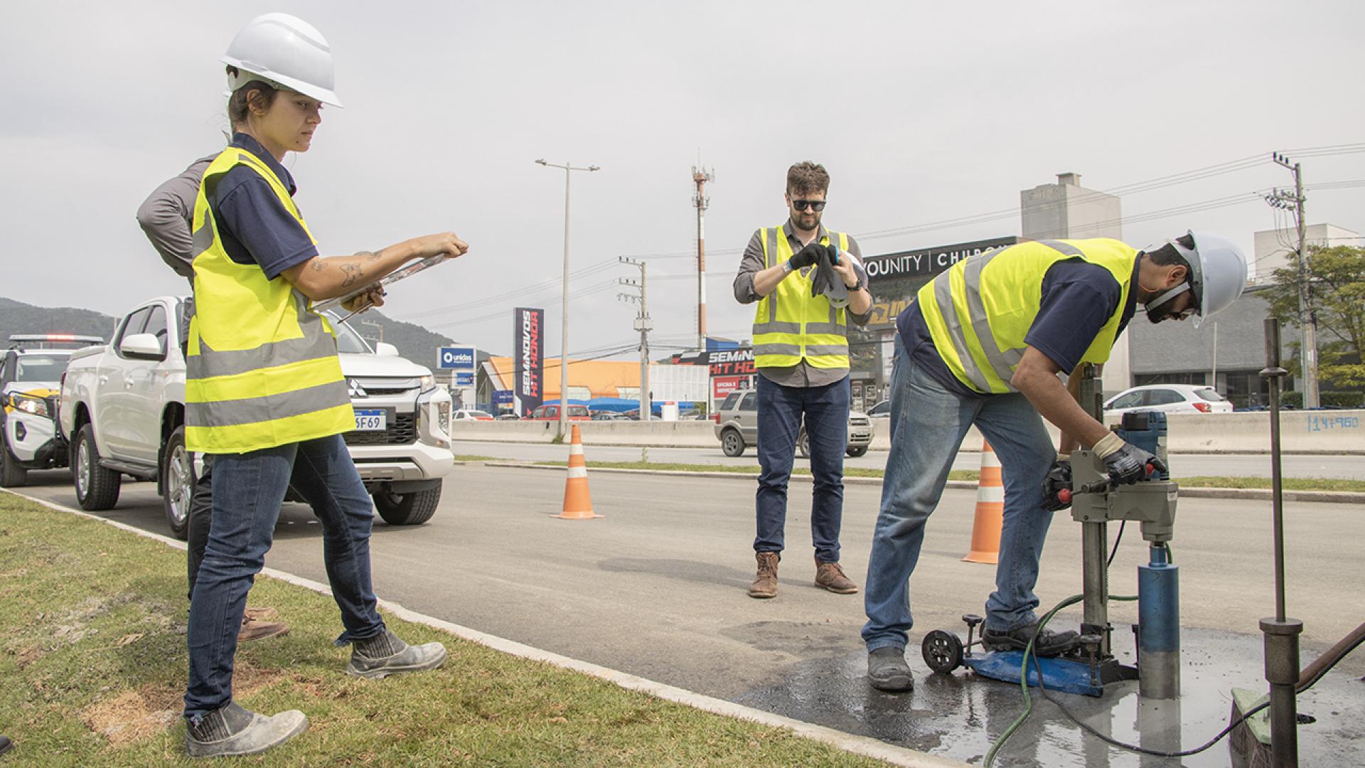 Foto mostra três pessoas em uma rodovia rajando coletes amarelos. A pessoa à esquerda está com uma planilha na mão e de capacete branco. A pessoa ao centro está de óculos escuros e calçando uma luva. A terceira, à direita, também usa capacete e manuseia uma máquina que fura o chão