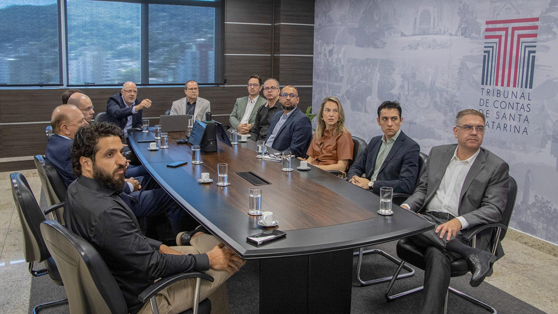 A imagem em formato horizontal apresenta 12 pessoas sentadas, sendo 11 homens e uma mulher, ela sentada na parte direita, e o grupo está observando uma apresentação. Ao fundo, na parede do lado direito está o logotipo do Tribunal de Contas de Santa Catarina