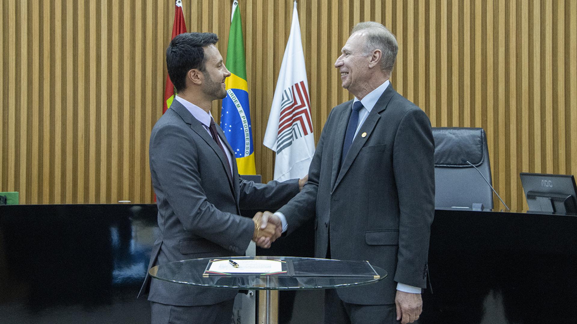 Foto do novo procurador Leandro Ocaña Vieira cumprimentando o presidente  do TCE/SC, conselheiro Herneus De Nadal. Ao fundo as bandeiras do Brasil, ao centro, do TCE/SC, à direita, e a de Santa Catarina à esquerda.