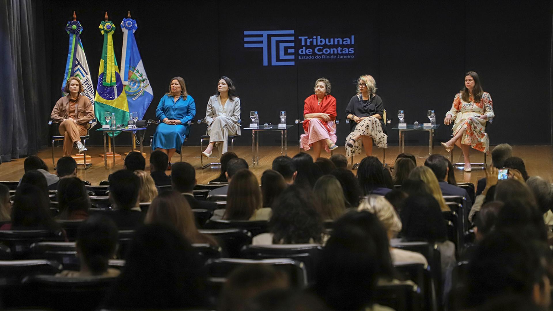 Banner horizontal com foto da plateia acompanhando um painel de debates realizado no TCE-RJ. No palco, seis mulheres estão sentadas em poltronas dispostas lado a lado, com mesas de apoio e copos de água à frente. Entre as palestrantes, está a conselheira substituta do TCE/SC Sabrina Iocken. 