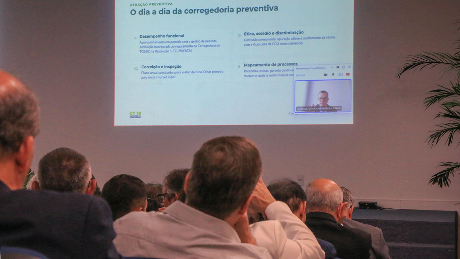 Banner horizontal com foto, em primeiro plano, da plateia assistindo à videoconferência com o conselheiro Adircélio, exibida no telão ao fundo. 