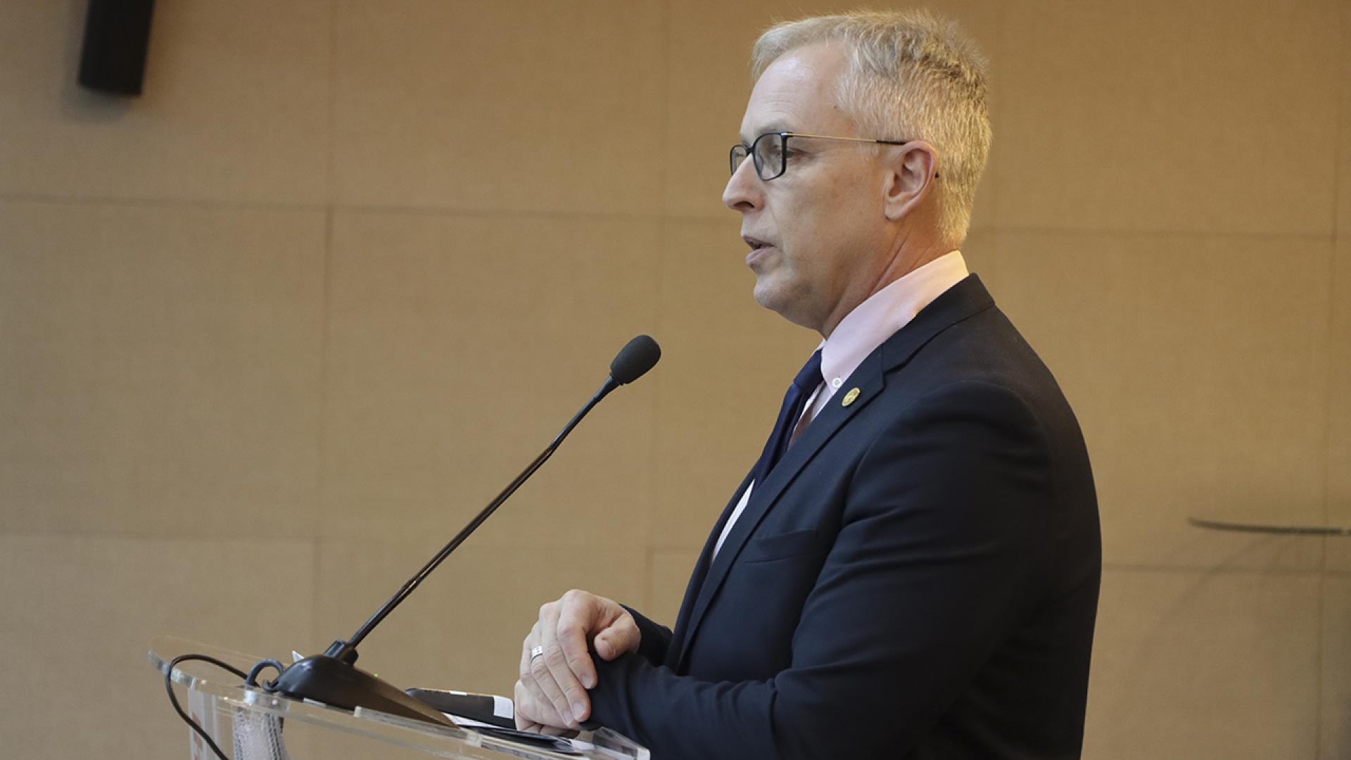 Banner horizontal com foto de perfil do diretor-geral de Controle Externo, Sidney Tavares, falando ao microfone durante sessão plenária. É um homem branco, de cabelos grisalhos. Veste terno e gravata e usa óculos de grau.