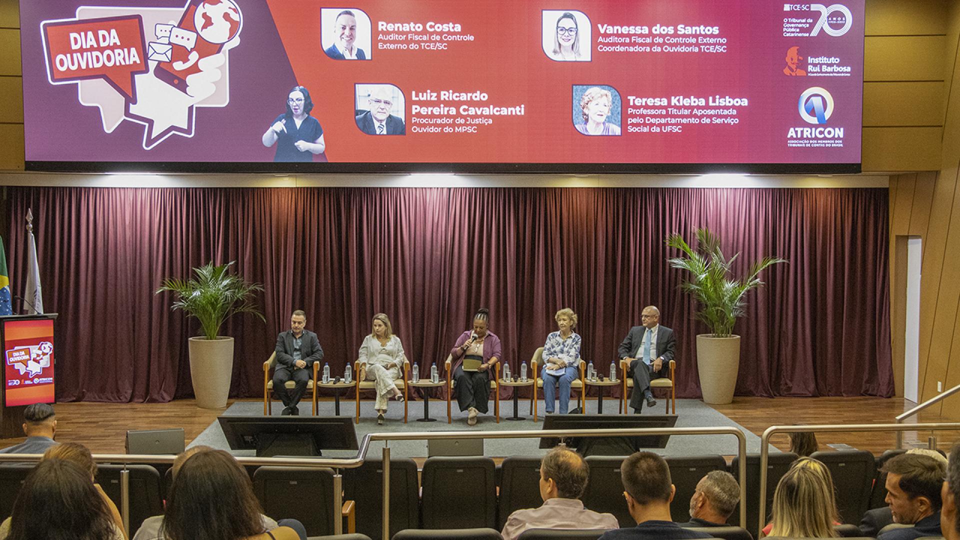 Foto do palco do TCE/SC, com os quatro palestrantes e ao centro, a diretora da Ouvidoria do TCE/SC, Walkiria Machado Rodrigues Maciel. ao fundo, o banner do evento.