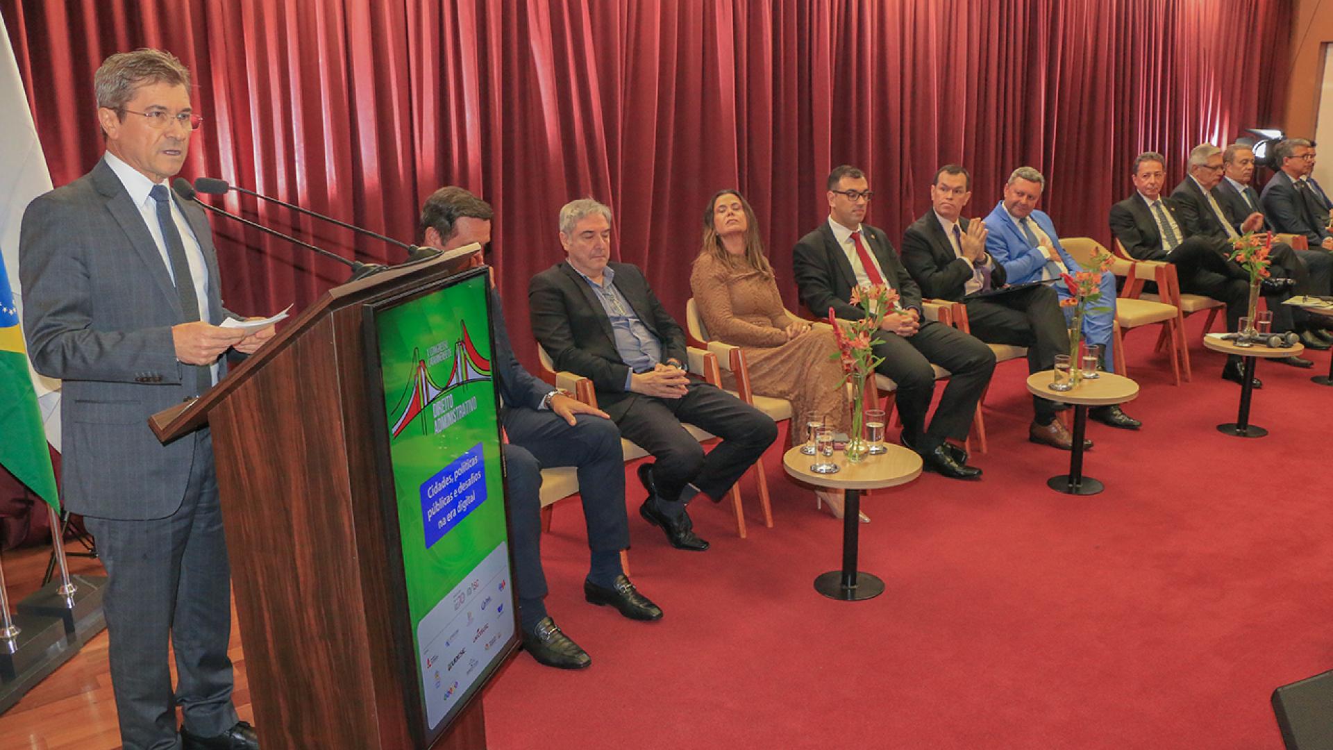 Banner horizontal com foto das autoridades no palco do auditório do TCE/SC durante a abertura do evento. Entre os presentes, os conselheiros José Nei Ascari e Adircélio Ferreira Júnior, o presidente do Idasc, José Sérgio Cristóvam, e a procuradora Cibelly Farias. Todas as autoridades estão lado a lado, sentadas em poltronas. No púlpito, discursa o conselheiro Ascari. O ambiente tem tapete e cortinas bordôs.