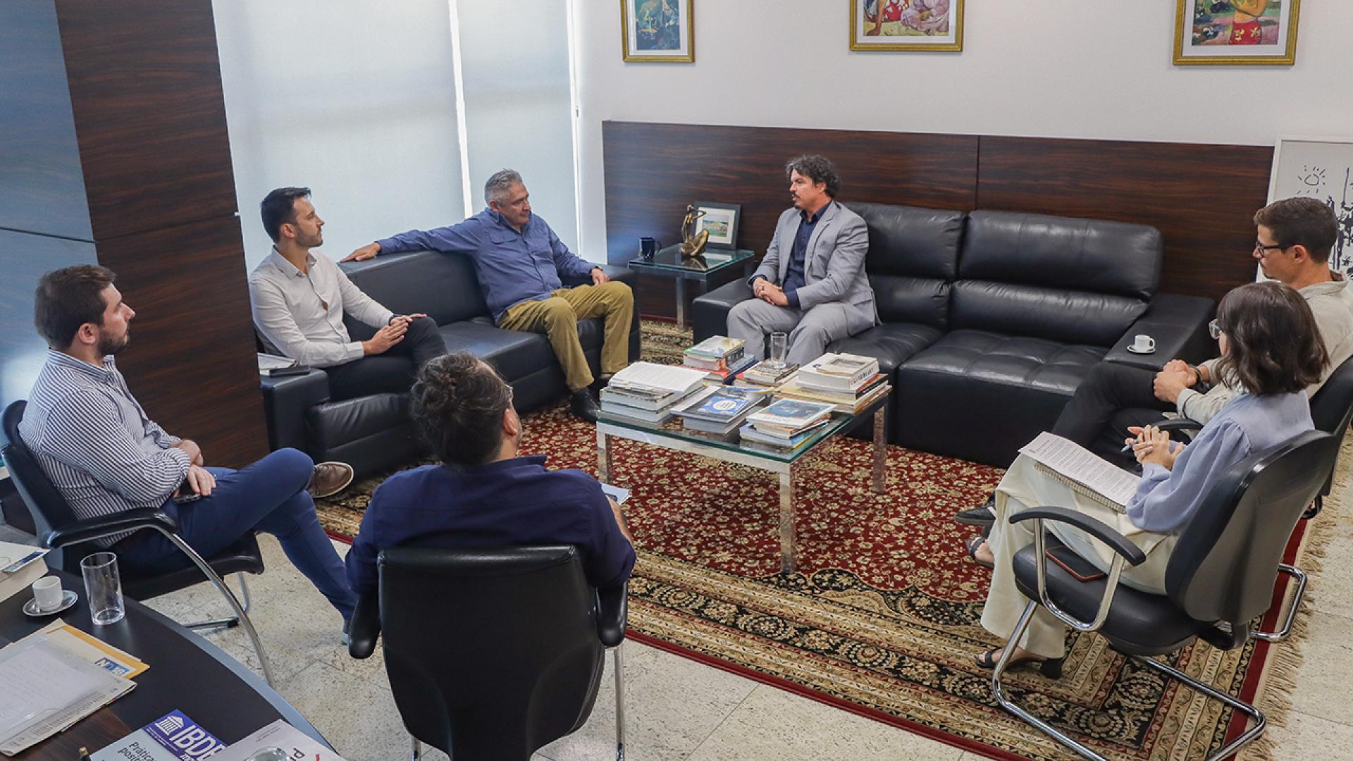 Imagem mostra sete pessoas sentadas em círculo, em sofás e cadeiras, numa reunião.
