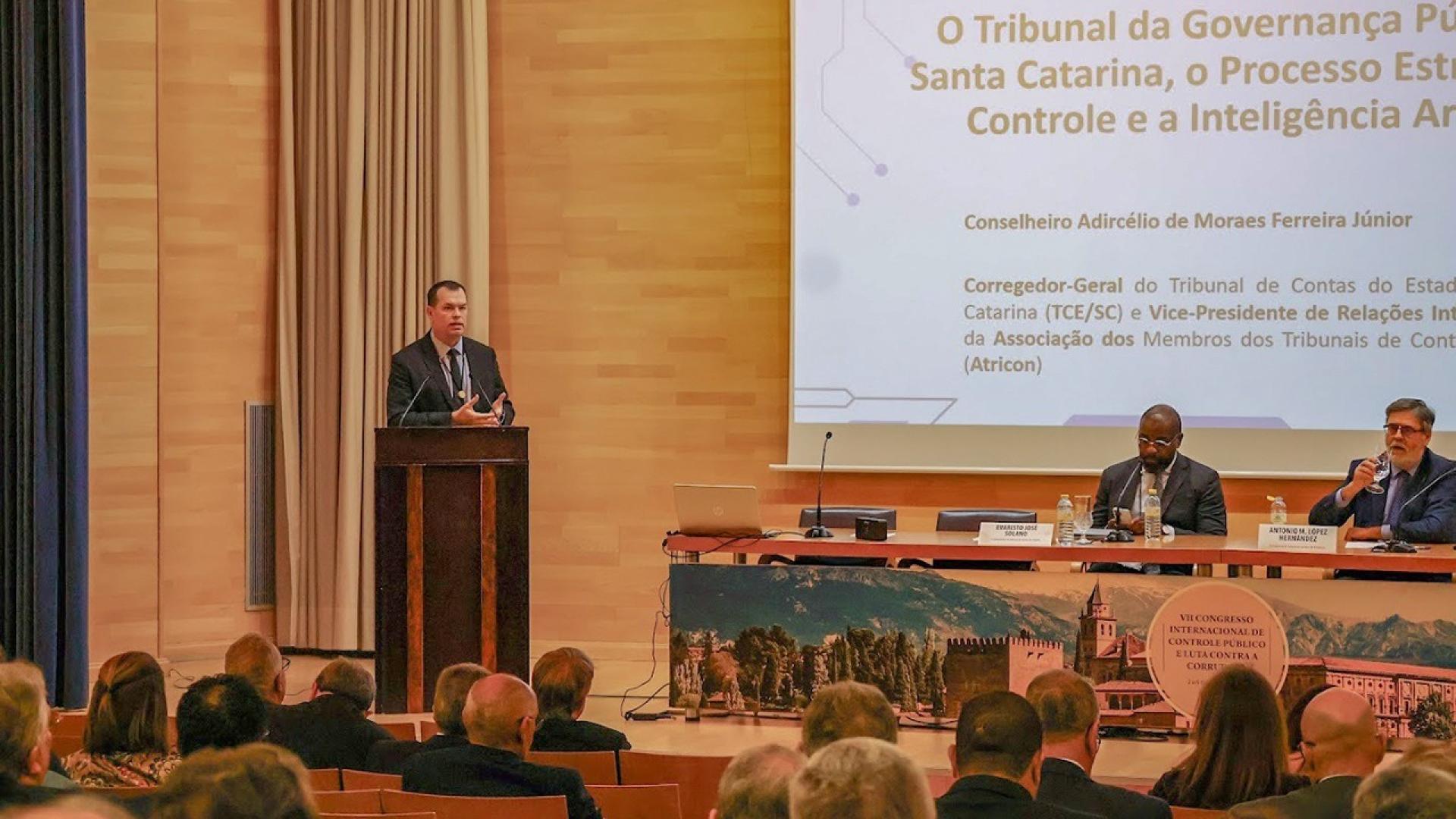 Banner horizontal com foto do conselheiro Adircélio de Moraes Ferreira Júnior, em pé, discursando no púlpito, no palco do auditório, durante o evento internacional.  À direita, mesa de autoridades. Ao fundo, telão com a apresentação. Em primeiro plano, à frente, pessoas de costas, sentadas, acompanhando a fala do conselheiro.  