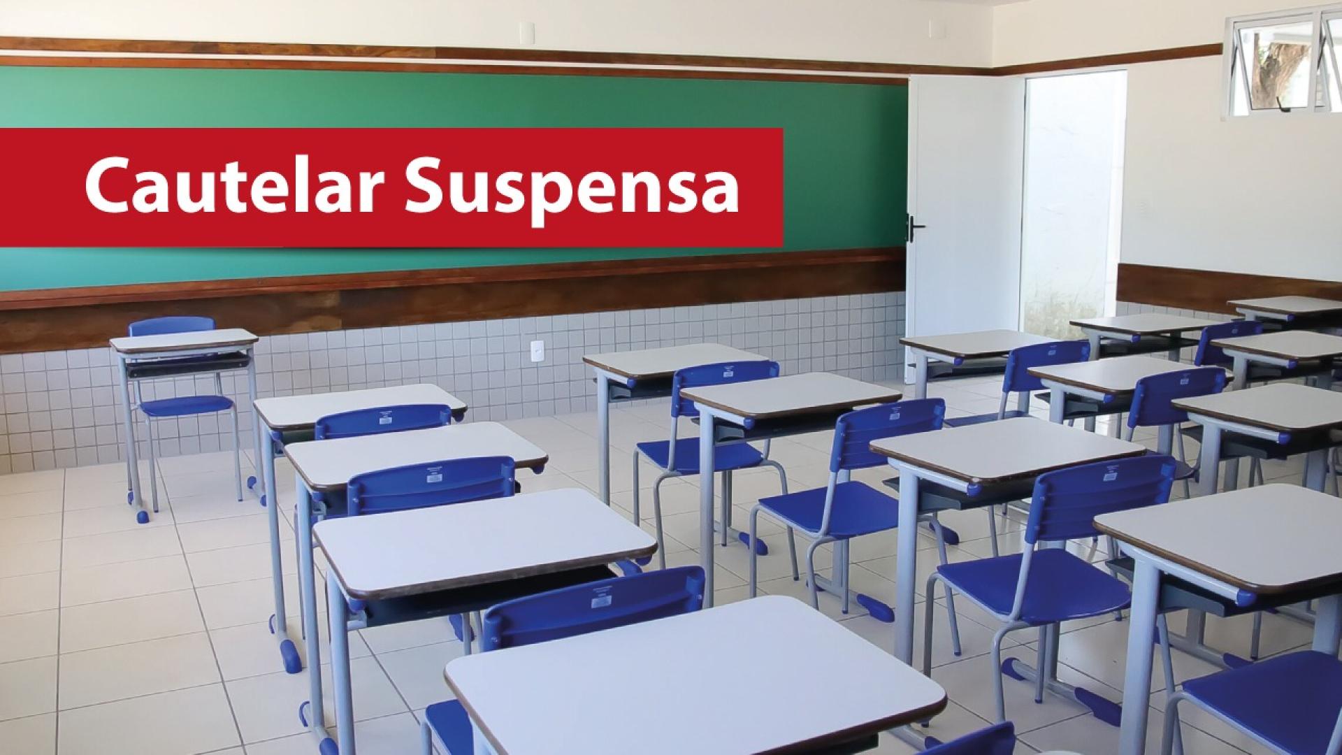 Banner horizontal com imagem de uma sala de aula com carteiras enfileiradas e vazias. Ao fundo, um quadro de giz. Sobre a foto, em tarja vermelha, o texto "Cautelar suspensa". 