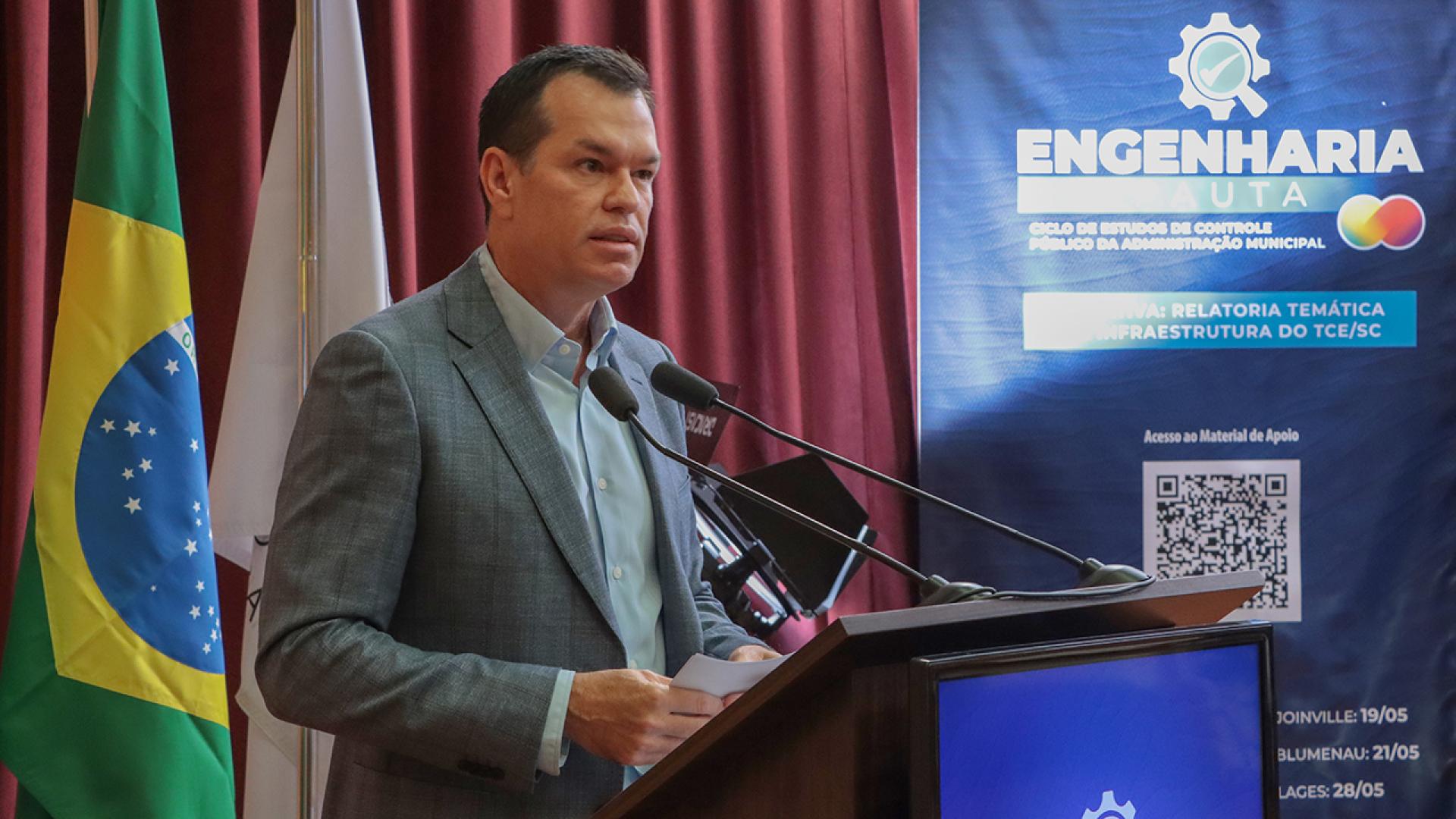 Banner horizontal com foto do conselheiro-corregedor, Adircélio de Moraes Ferreira Júnior, falando ao púlpito, no auditório do TCE/SC. Ele é um homem branco, de cabelos escuros. Veste paletó cinza. Ao seu lado, banner vertical com logo do Engenharia em Pauta. Ao fundo, cortinas bordôs.