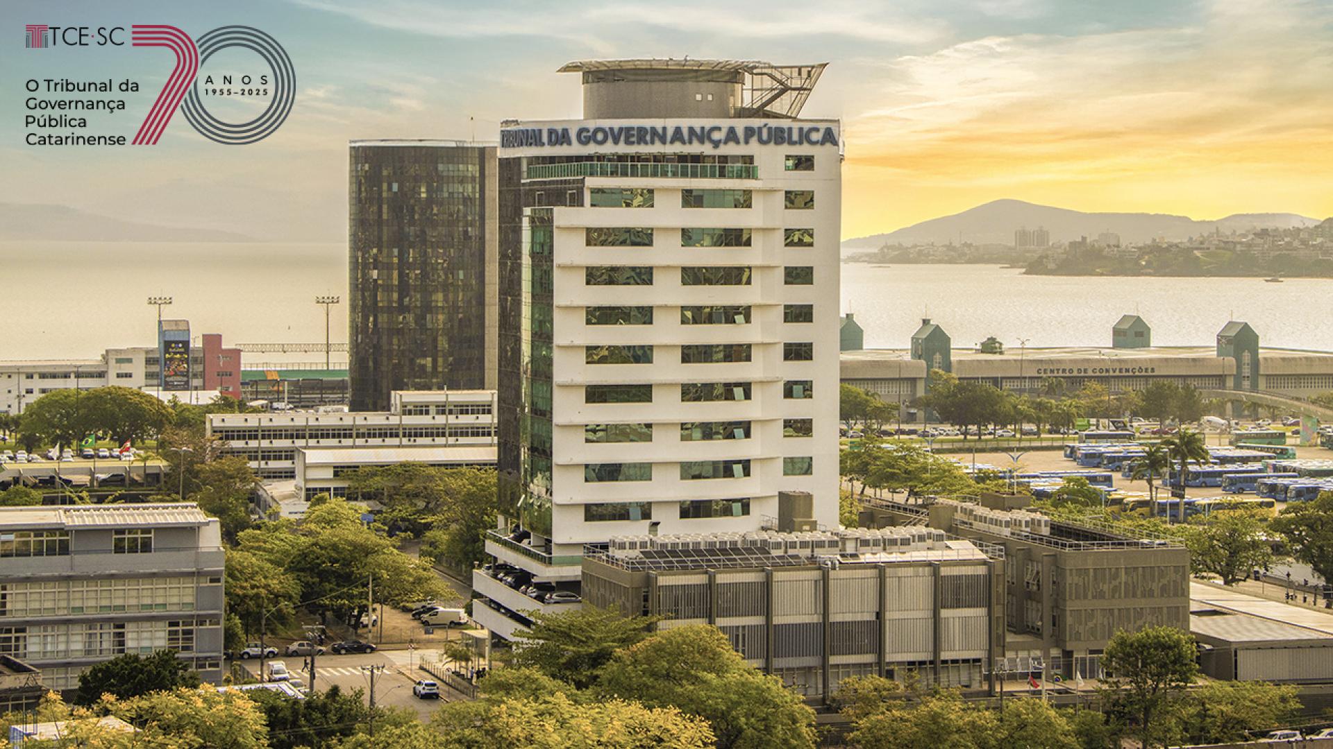 Banner horizontal com foto aérea do edifício do TCE/SC ao entardecer. A sede é um prédio de 13 andares de vidro espelhado e na cor branca. Na fachada, há o texto "Tribunal da Governança Pública". Compõem a paisagem, ao fundo, o mar, árvores e demais construções. Sobre a foto, no canto superior esquerdo, selo comemorativo aos 70 anos do TCE/SC.  