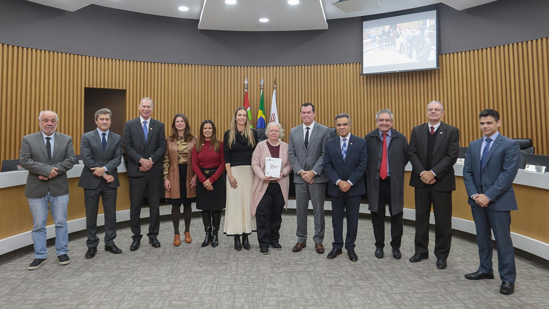 Banner horizontal com foto de 12 pessoas em pé, no Plenário do TCE/SC, durante entrega dos exemplares a Hanne Kristoffersen. Entre os presentes, além da bibliotecária, o presidente, o vice-presidente e o corregedor-geral do TCE/SC, conselheiros Herneus De Nadal, José Nei Ascari e Adircélio Ferreira Júnior, o presidente da Atricon, Edilson Silva, e a integrante da equipe executiva da revista, Juliana Fritzen. 