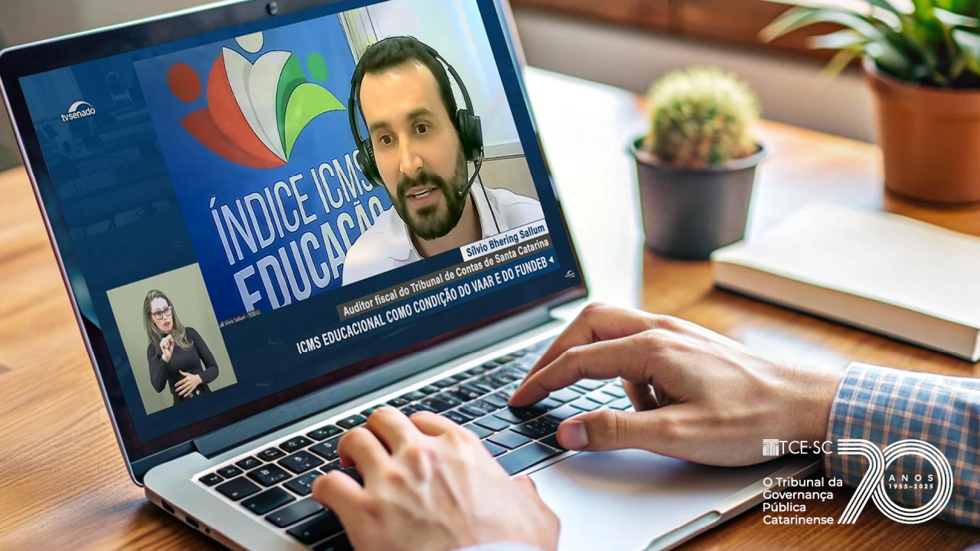 foto de um notebook aberto e aparecem as mãos de uma pessoa branca digitando Na tela do computador aparece a imagem do servidor do TCE/SC Silvio Sallum, durante audiência sobre ICMS Educação no Senado Federal
