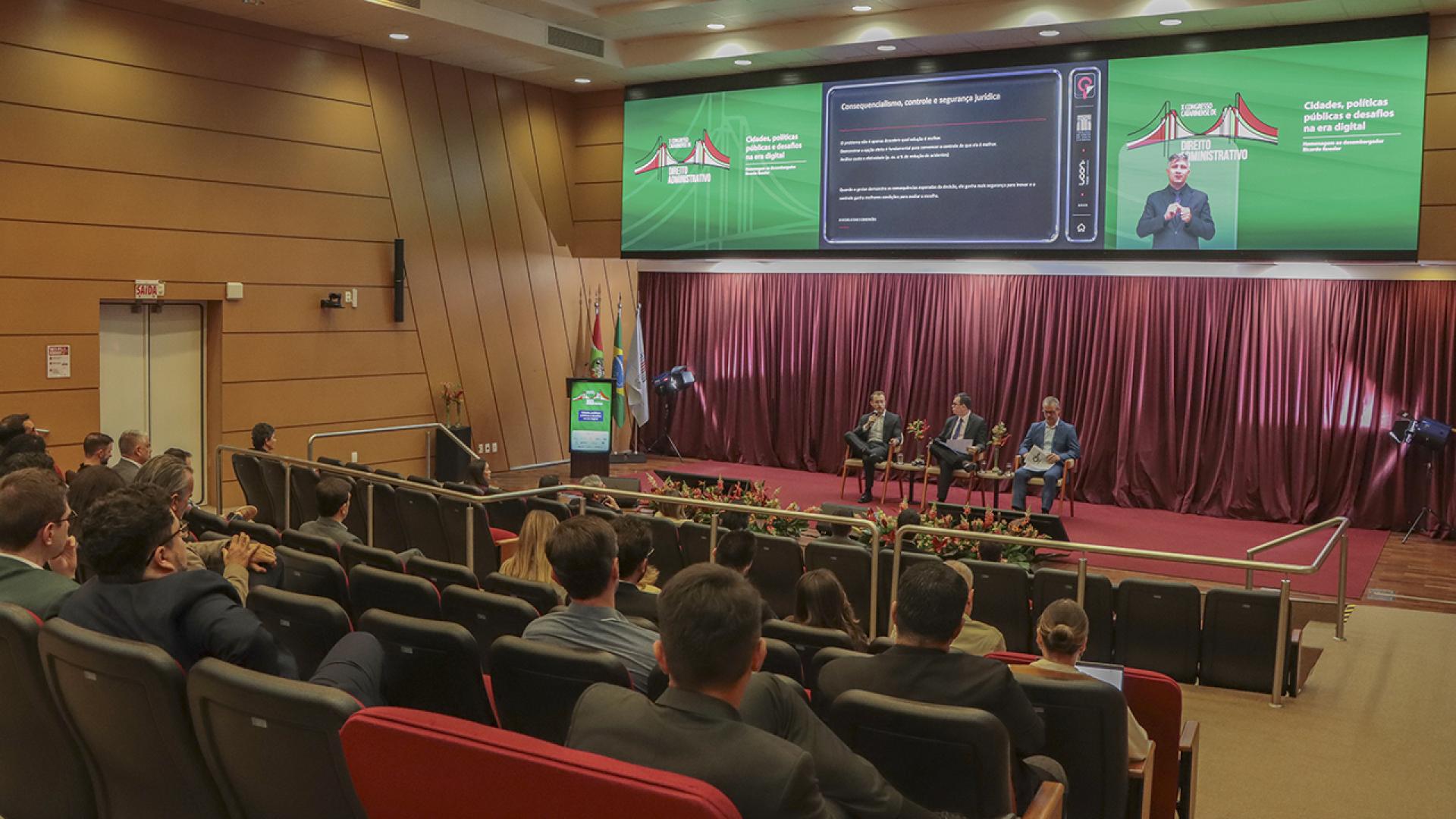 Foto horizontal do auditório vermelho do TCE/SC. No placo os intregantes do painel 1, entre eles o conselheiro do TCE/SC Adircélio de Moraes Ferreira Júnior. Ao fundo do palco, sobre a cortina, há o telão com a logo do evento 
