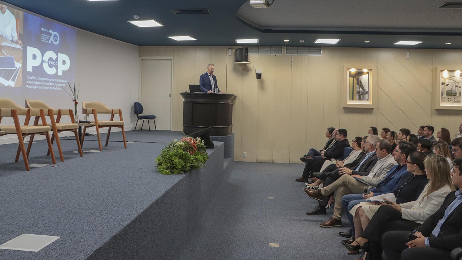 Banner horizontal com foto de um auditório durante um evento institucional. Em destaque, o presidente Herneus está em pé, atrás de um púlpito sobre o palco, enquanto o público acompanha a apresentação sentado em fileiras à direita da imagem. No telão ao fundo aparecem a identidade visual do evento e a sigla “PCP”. O palco conta com poltronas para debatedores