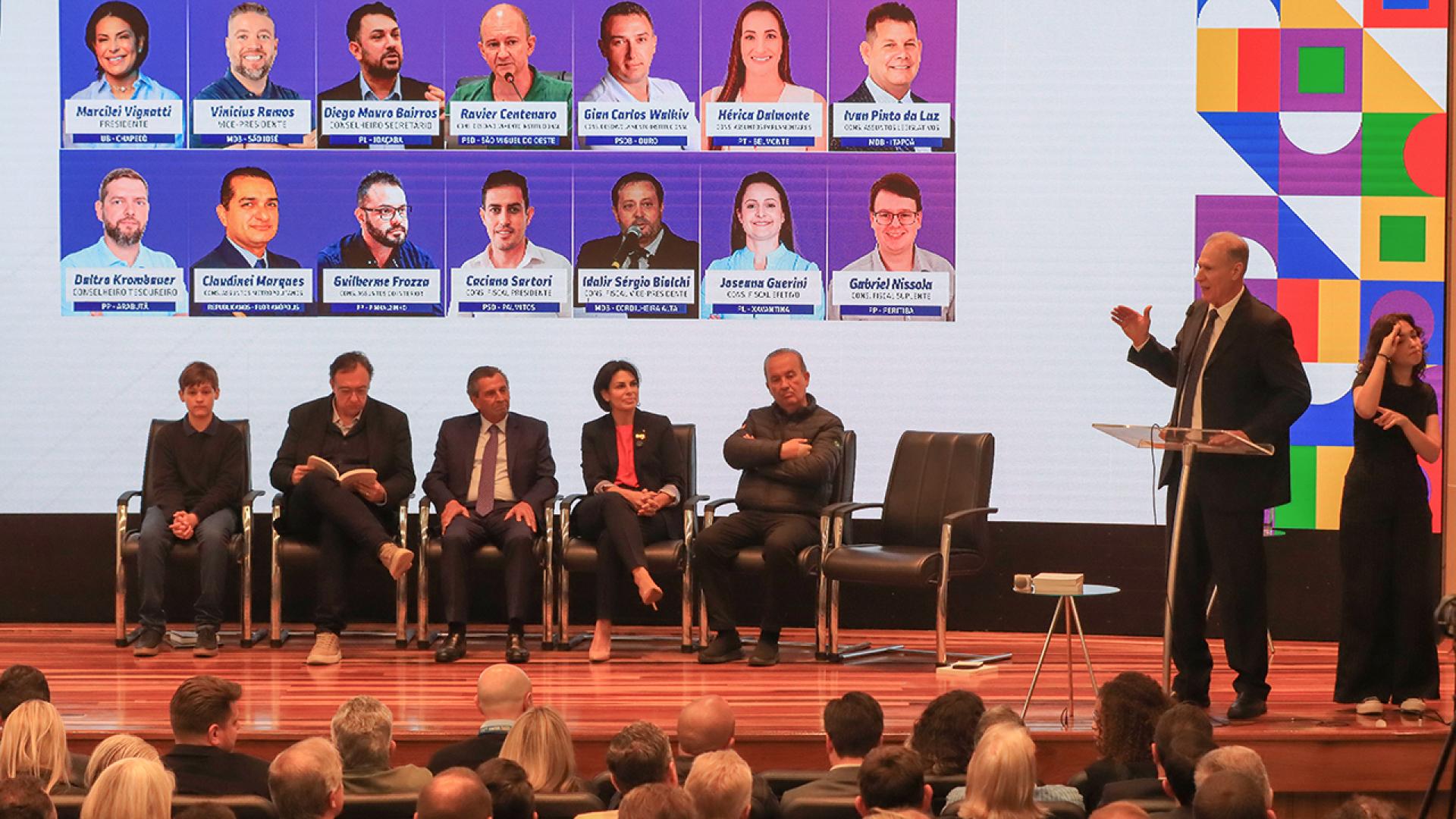 Foto do palco do auditório da Alesc. O presidente do TCE/SC, conselheiro Herneus De Nadal, está no púlpito, ao lado direito da foto, fazendo sua manifestação na posse da diretoria da Uvesc. No palco, estão sentados, da esquerda para a direita, o vereador mirim, João Pedro Probst, a nova presidente da Uvesc,  Marcilei Vignatti; Gilson Conzatti, presidente da UVB; o presidente da Alesc, Julio Garcia,; a presidente da UVESC, Marcilei Vignatti; e o governador Jorginho Mello.