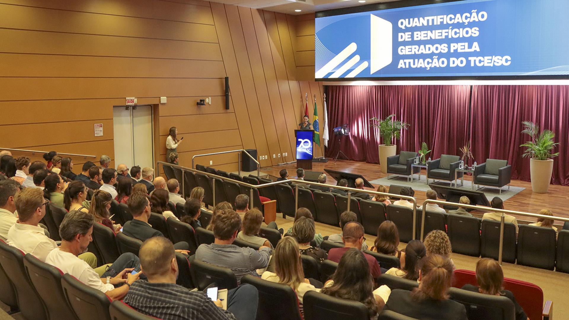 Banner horizontal com foto dos participantes do evento de treinamento no auditório do TCE/SC, entre diretores, servidores e autoridades do Tribunal, acomodados em poltronas enquanto acompanham a apresentação no palco. Eles estão de costas. Ao fundo, o púlpito, as bandeiras institucionais e o painel com o tema “Quantificação de benefícios gerados pela atuação do TCE/SC”, em ambiente formal e organizado.