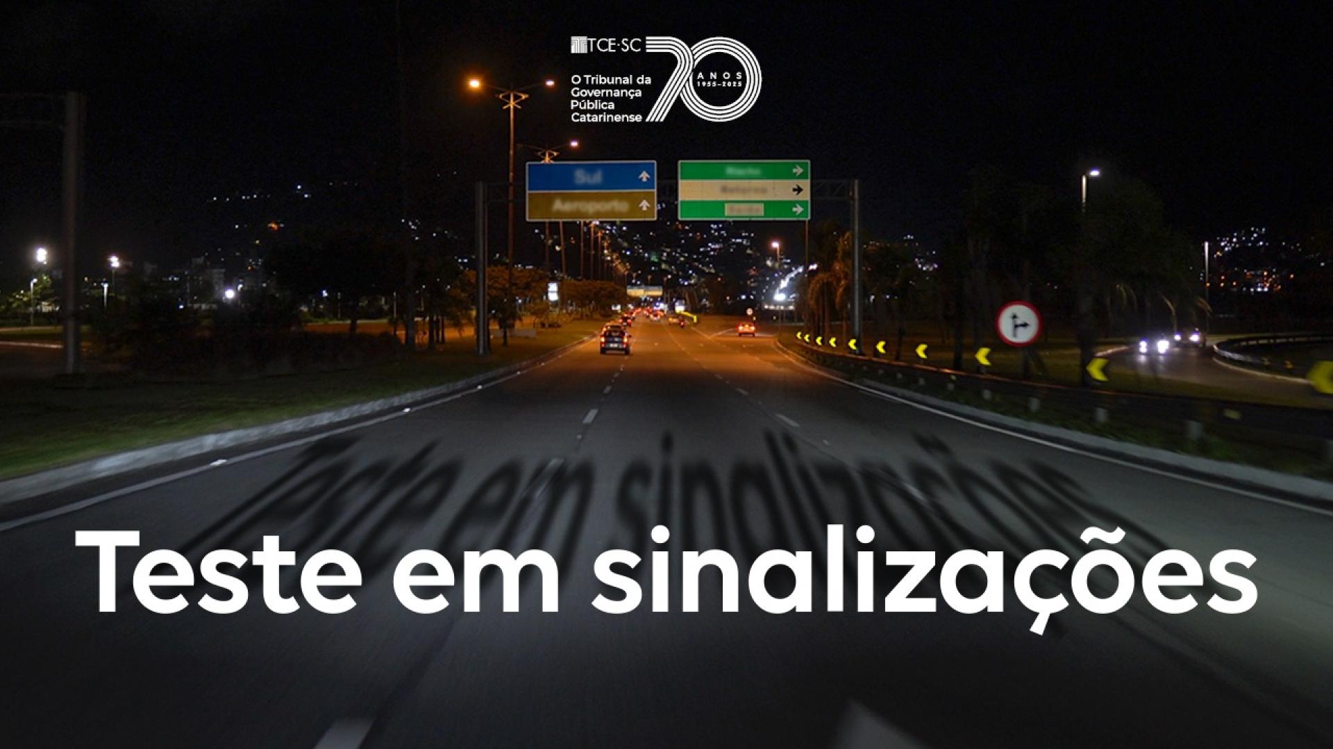 Imagem feita á noite mostra uma via iluminada, com as luzes dos faróis iluminando as placas de indicação de trânsito. Ao centro, escrito em branco, está escrito “teste em sinalizações”. No alto, ao centro, está o logo de 70 anos do TCE/SC