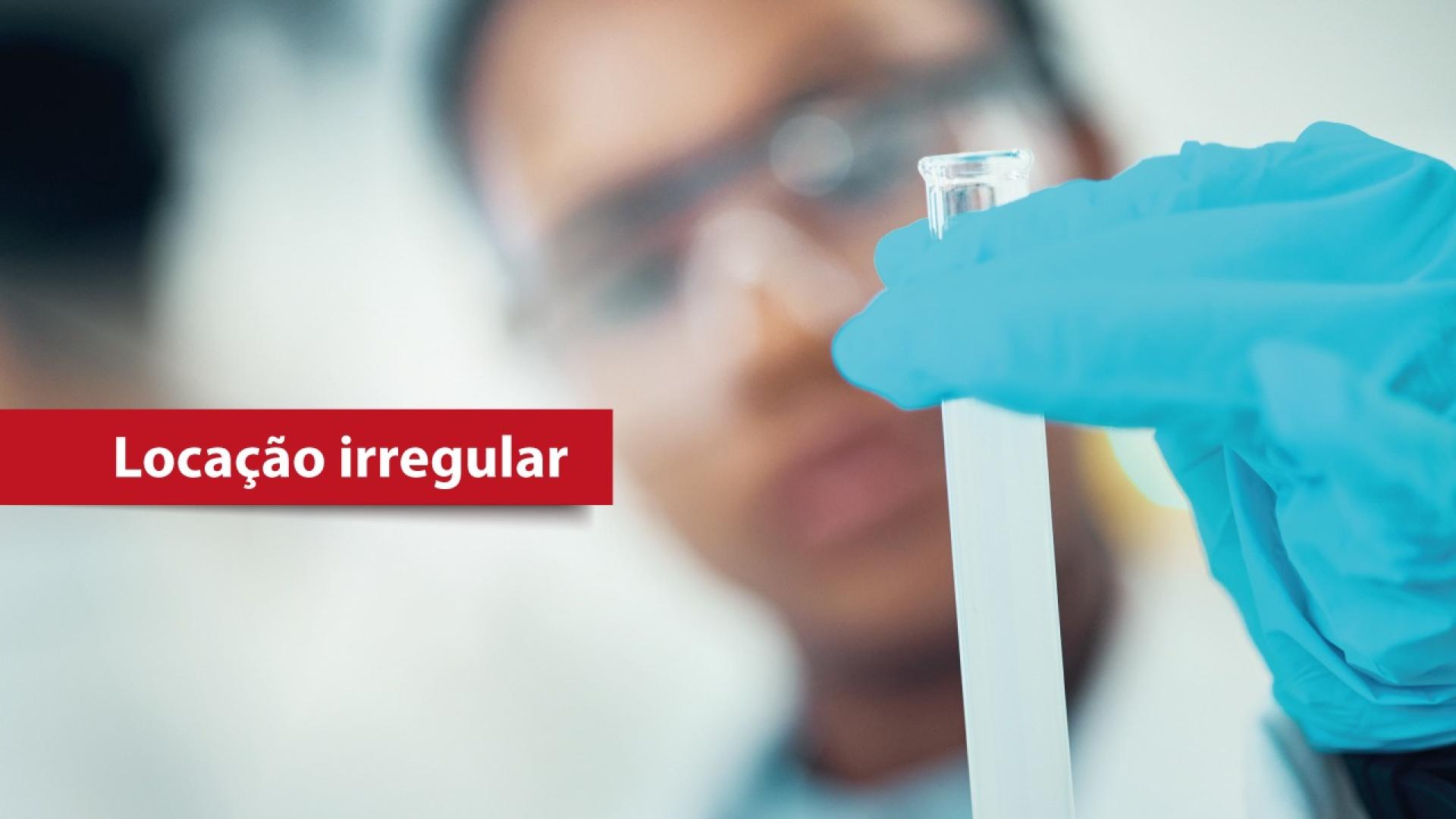 Banner horizontal que mostra, em primeiro plano, uma mão com luva azul segurando um tubo de ensaio com líquido claro. Ao fundo, desfocado, aparece uma pessoa usando óculos de proteção em ambiente que lembra um laboratório. À esquerda da imagem há uma faixa vermelha com o texto “Locação irregular”. 