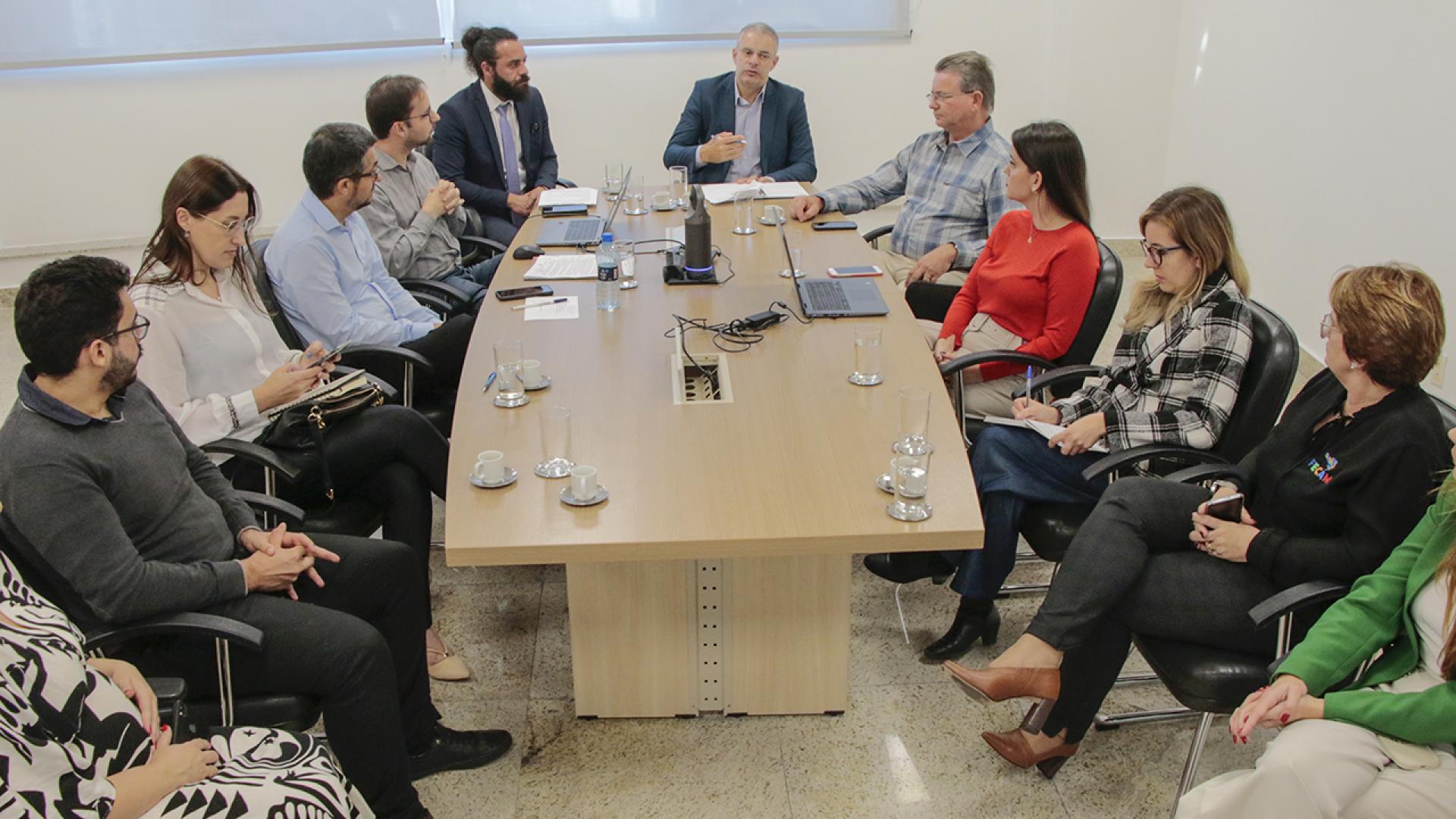 Foto de 11 pessoas sentadas diante de uma mesa, durante reunião. Entre homens e mulheres, está o conselheiro Gerson dos Santos Sicca, ao centro, um homem branco, de cabelos grisalhos, que está vestindo terno azul e camisa de cor clara.  