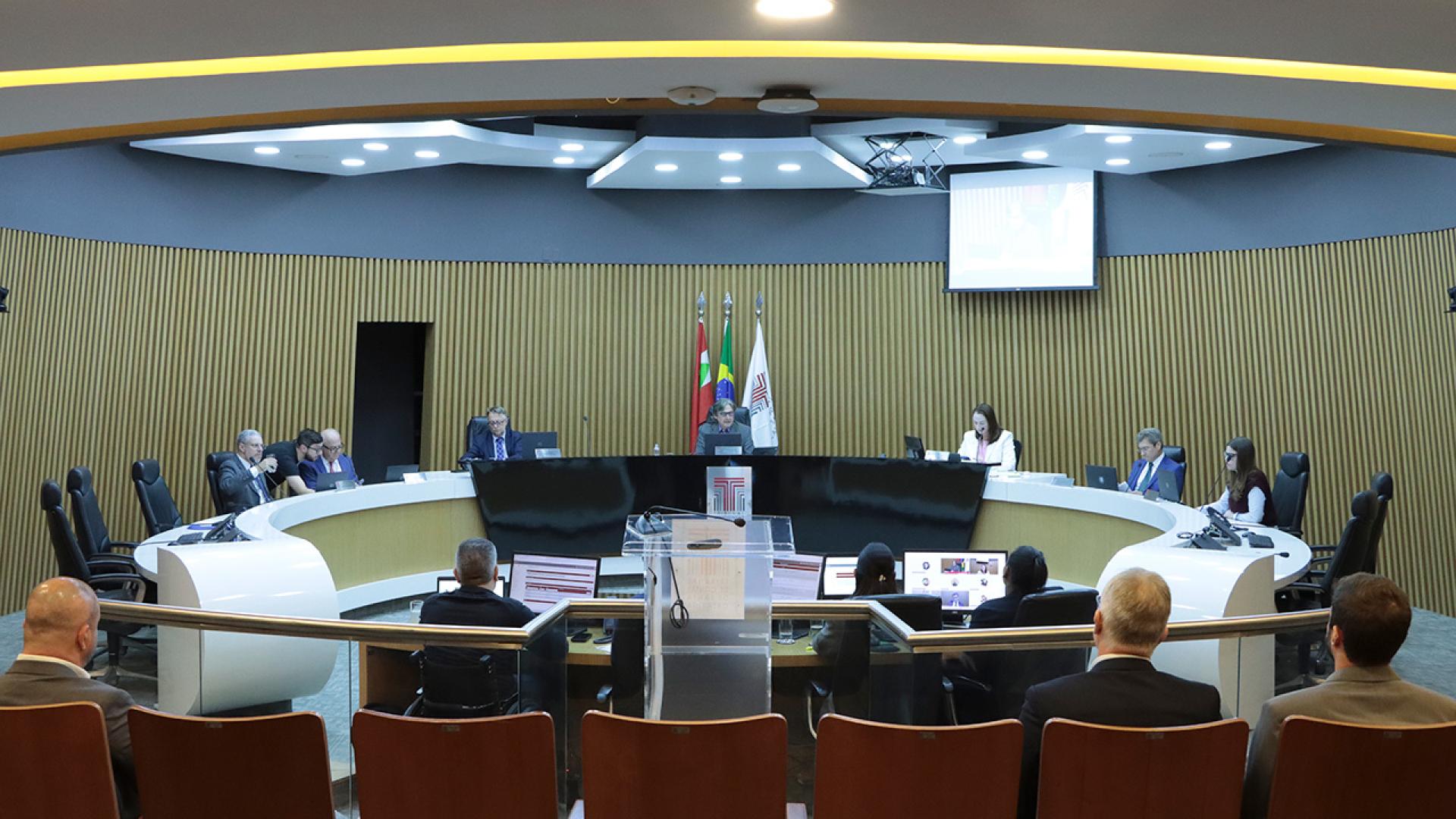 Banner horizontal com foto dos integrantes da Primeira Câmara no Plenário do TCE/SC. Ao centro da bancada circular, o presidente Aderson Flores. Ao redor, entre os presentes, os conselheiros Wilson Wan-Dall e José Nei Ascari, os conselheiros substitutos Gerson Sicca e Sabrina Iocken e o procurador Diogo Ringenberg. 