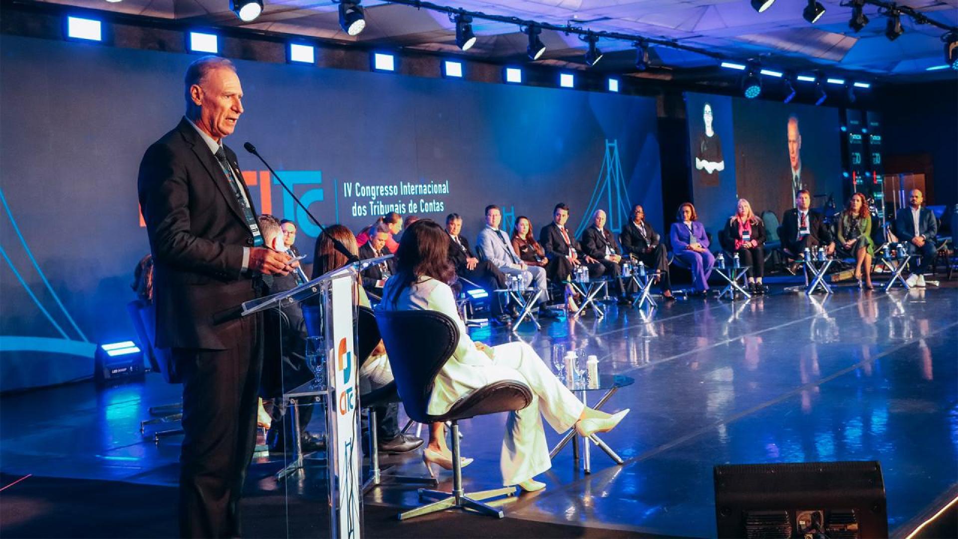 Foto com fundo azul. Em primeiro plano, à esquerda, homem trajando terno preto fala ao microfone. Ao fundo uma série de pessoas sentadas em perspectiva