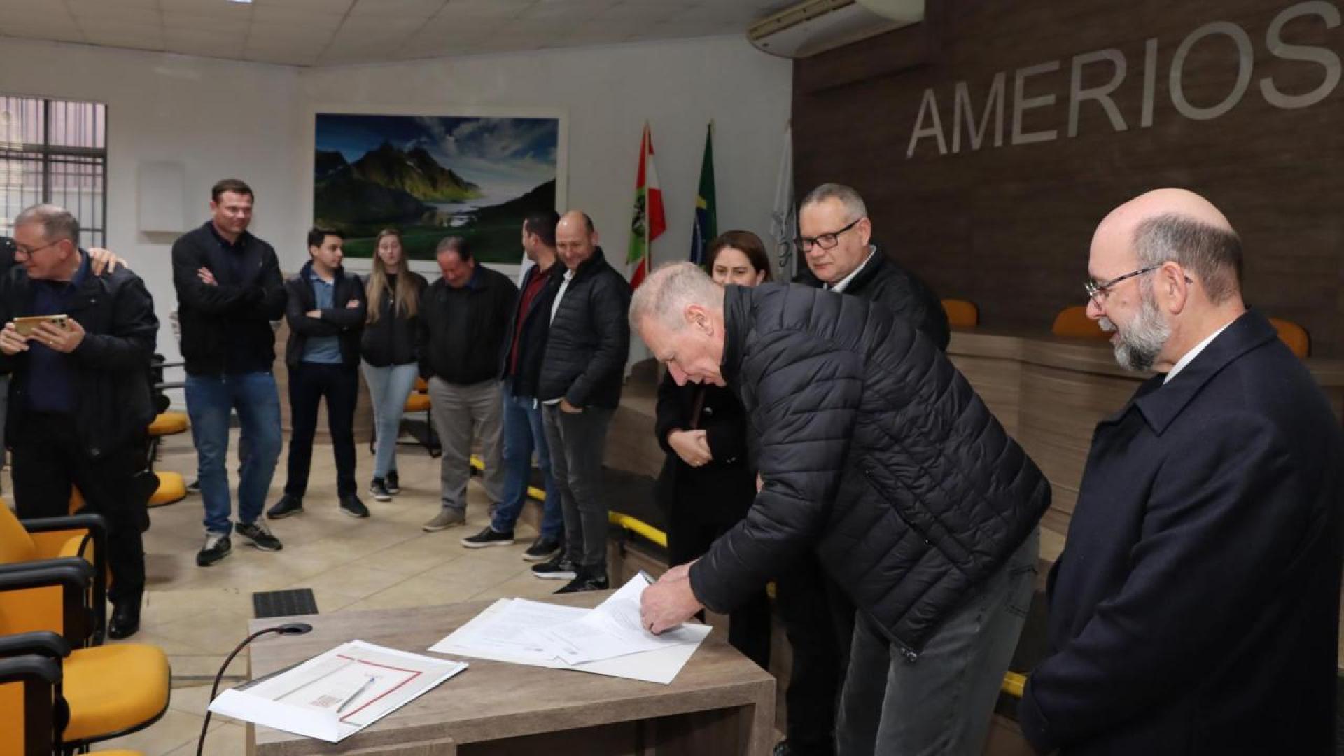 Banner horizontal com foto dos participantes do acordo em uma sala de reuniões da Amerios. No centro da imagem, o presidente do TCE/SC, conselheiro Herneus De Nadal, inclina-se sobre uma mesa para assinar o documento. Ao seu lado, acompanha o ato o assessor Moisés Hoegenn. Outras pessoas observam a assinatura ao redor da mesa. Ao fundo, aparecem a identificação “AMERIOS” na parede e bandeiras posicionadas no ambiente.  